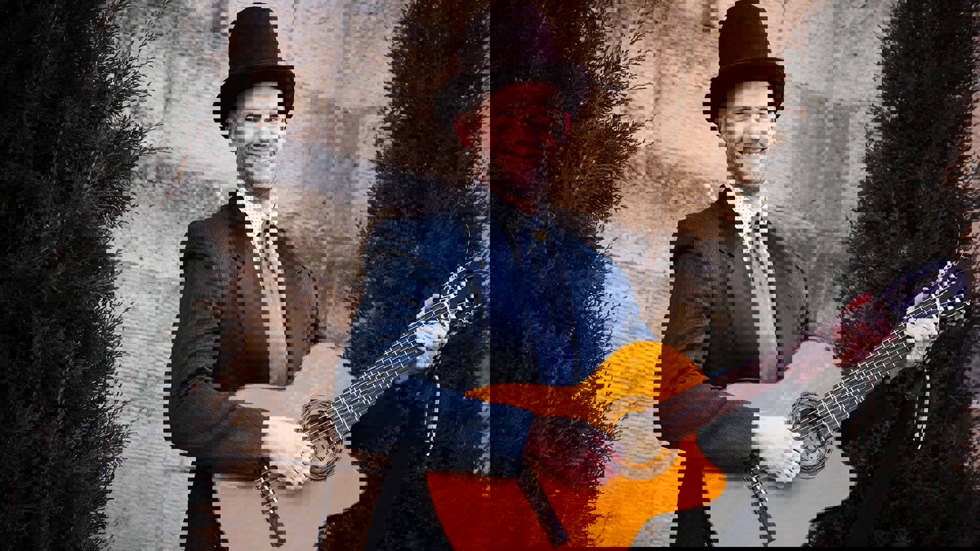 A white man in a navy suit and tophat holding an acoustic guitar. The background is a stone wall with two trees.