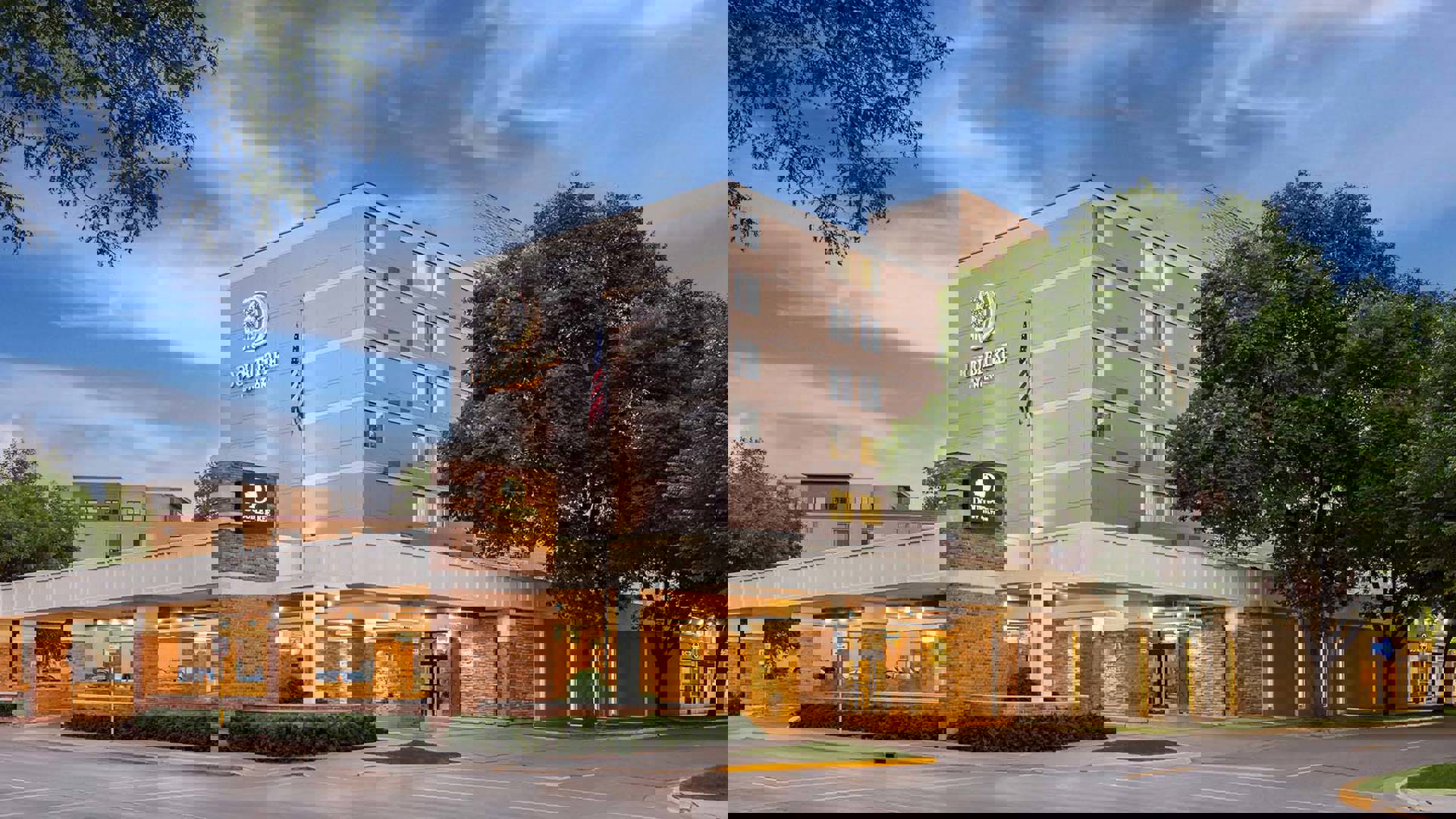 Image of a Double Tree Hotel exterior with a driveway and front entrance overhang. The building is about 5 stories tall, cream colored and flanked by trees.