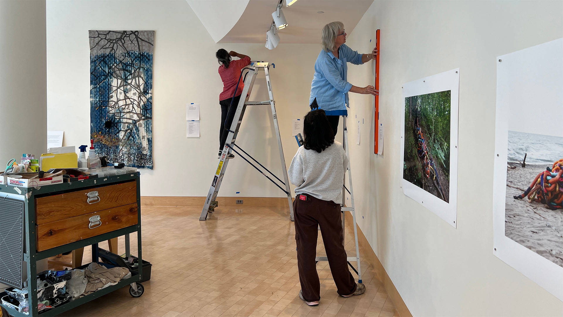 Three people installing pieces of art in a gallery with white walls.