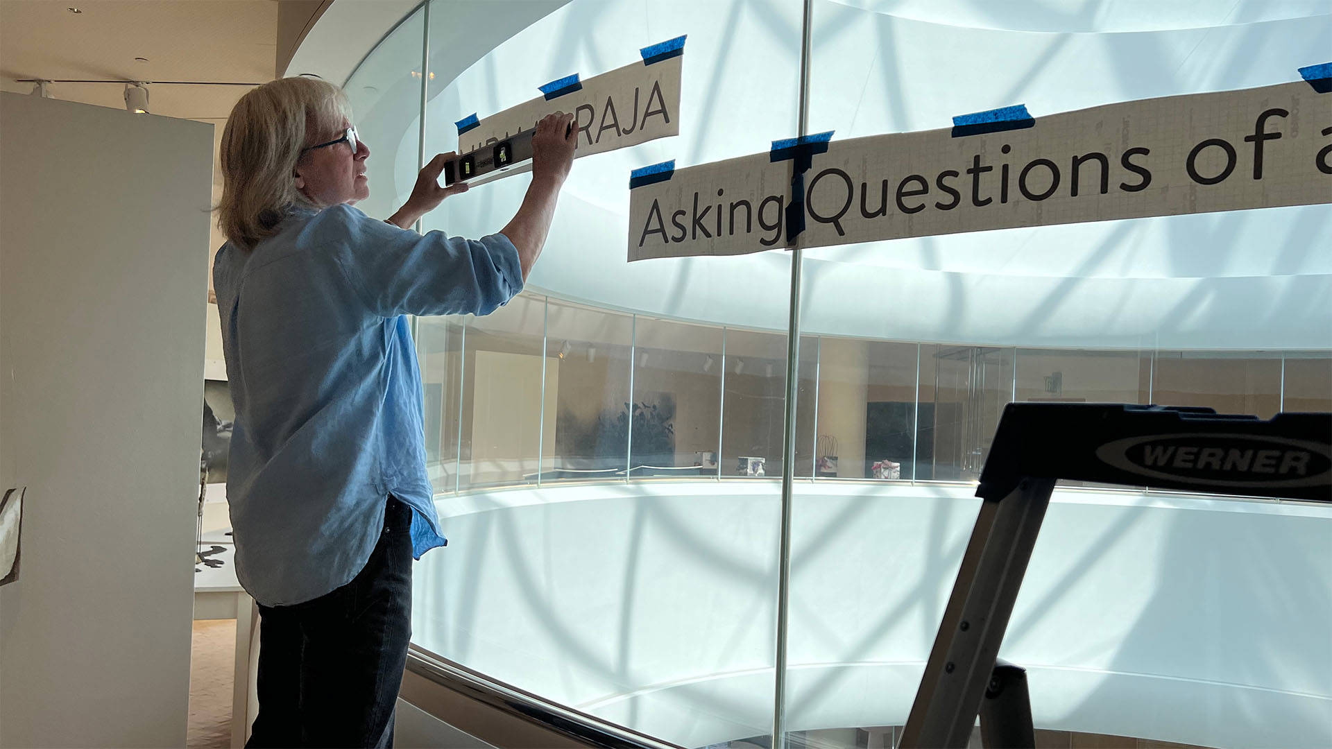 A middle aged white woman with short grey hair and glasses in a blue blouse using a level to check the placement of a sign on a large glass window.