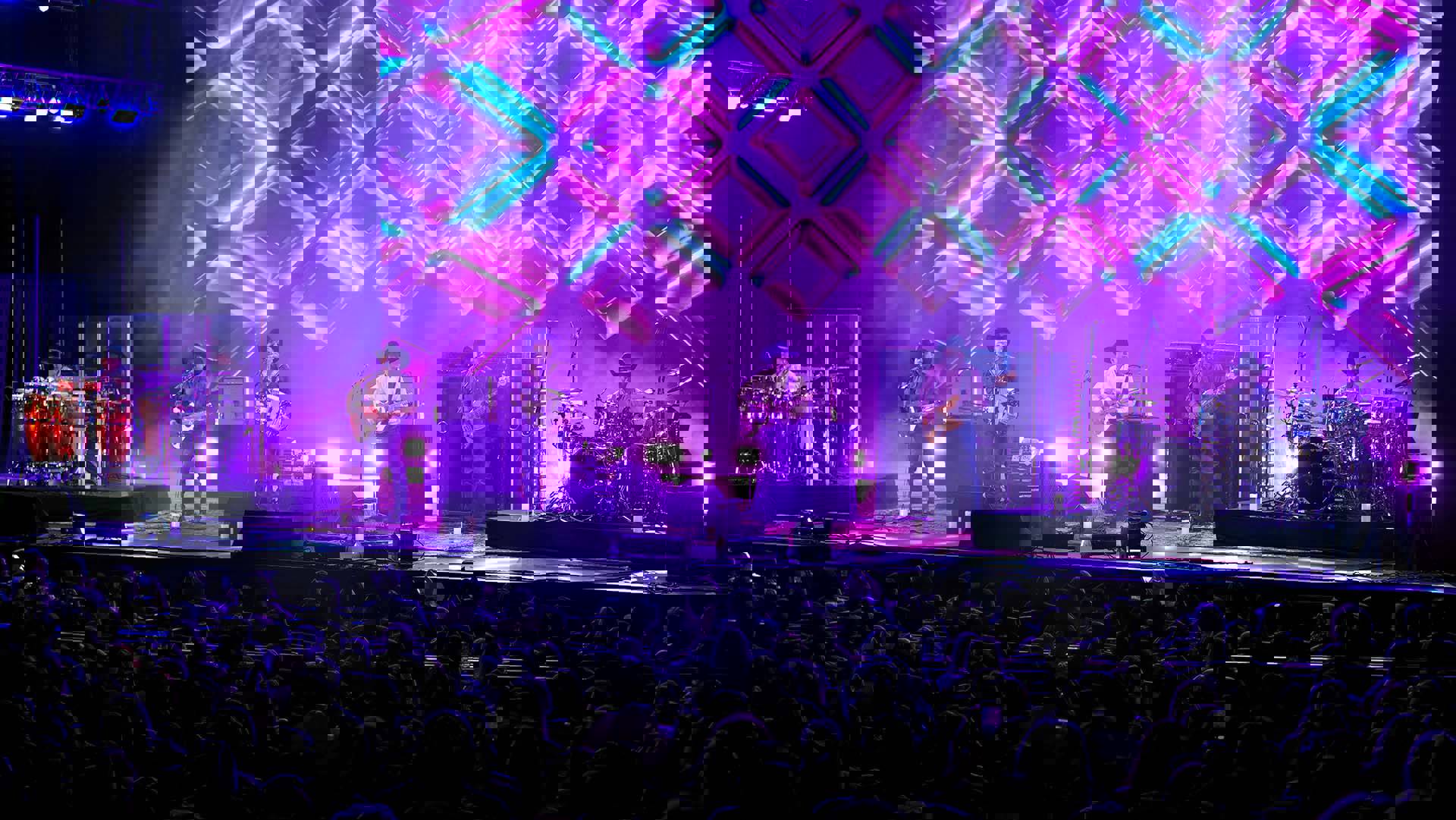 Intocable, a six-piece band performing on stage to a crowd. Bright purple and blue shapes are displayed on a large screen behind them.