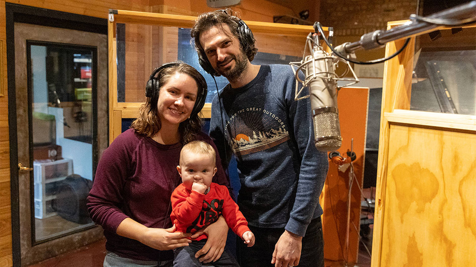 A caucasian woman and man holding an infant in an orange sweater standing in a recording booth with large headphones on.