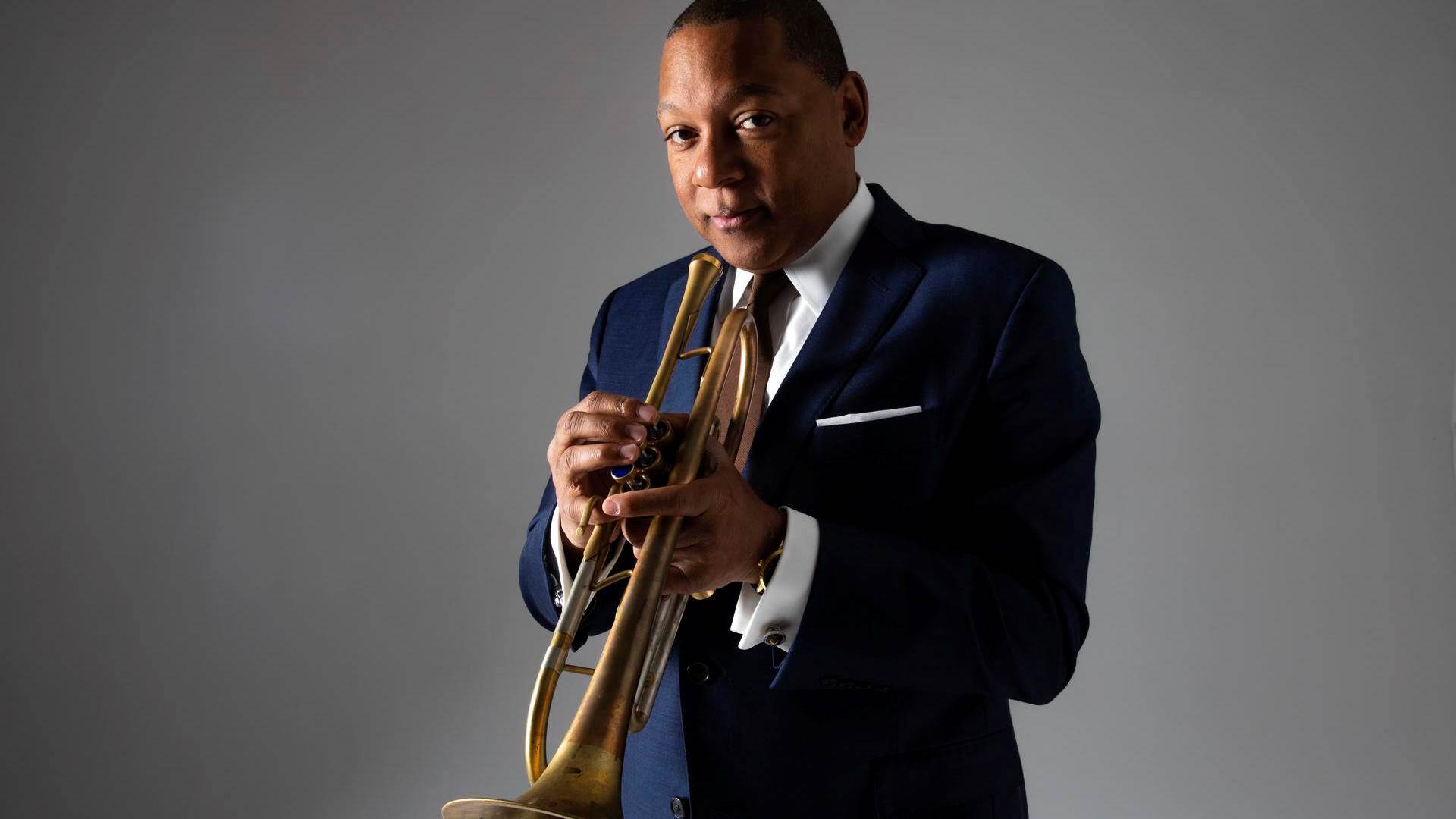Wynton Marsalis, a middle aged man with dark complexion and brown eyes in a blue suit and brown tie holding a trumpet. The background is grey.