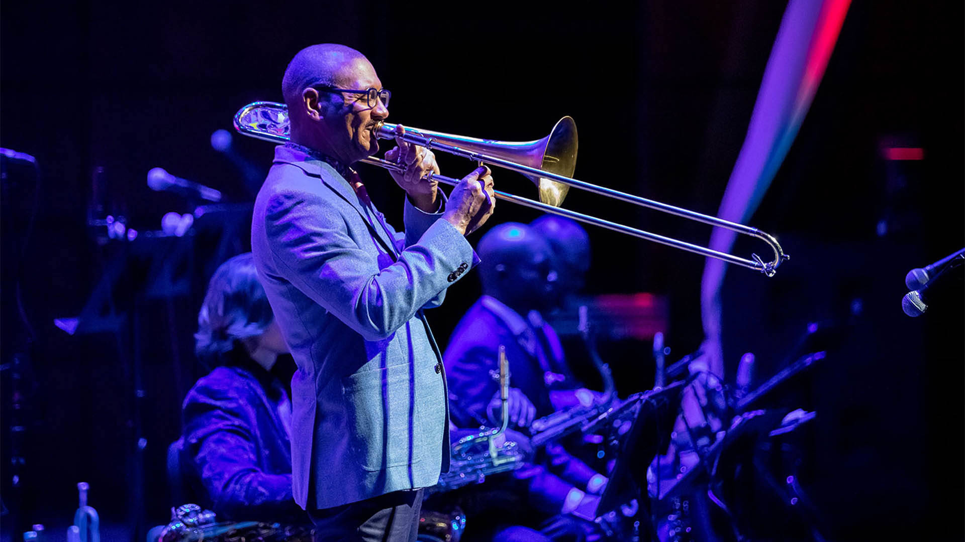 A Black man with buzzed hair and glasses wearing a blue suit and playing a trombone while standing.