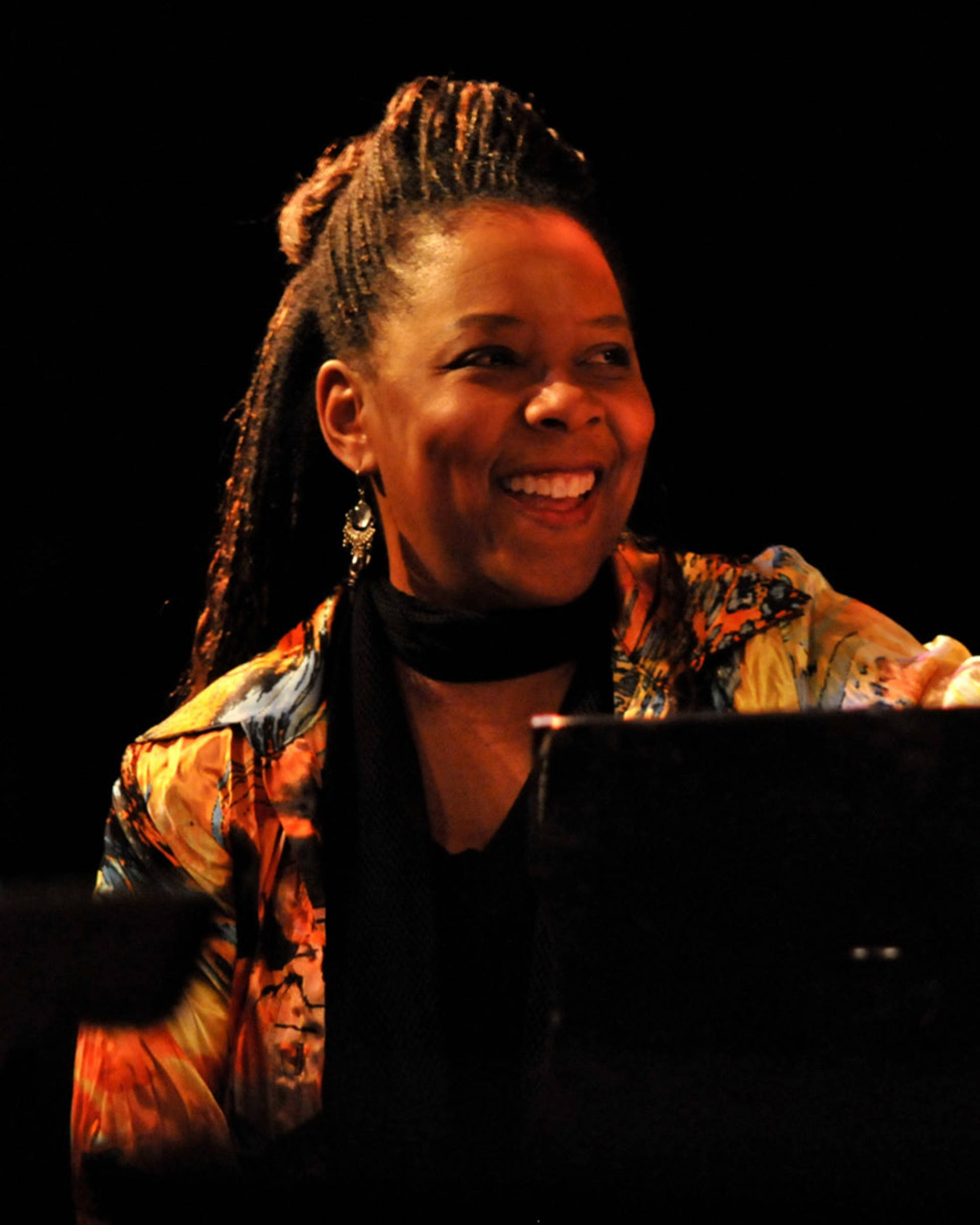 A smiling african american woman with long dark hair and a colorful top sitting at a piano.