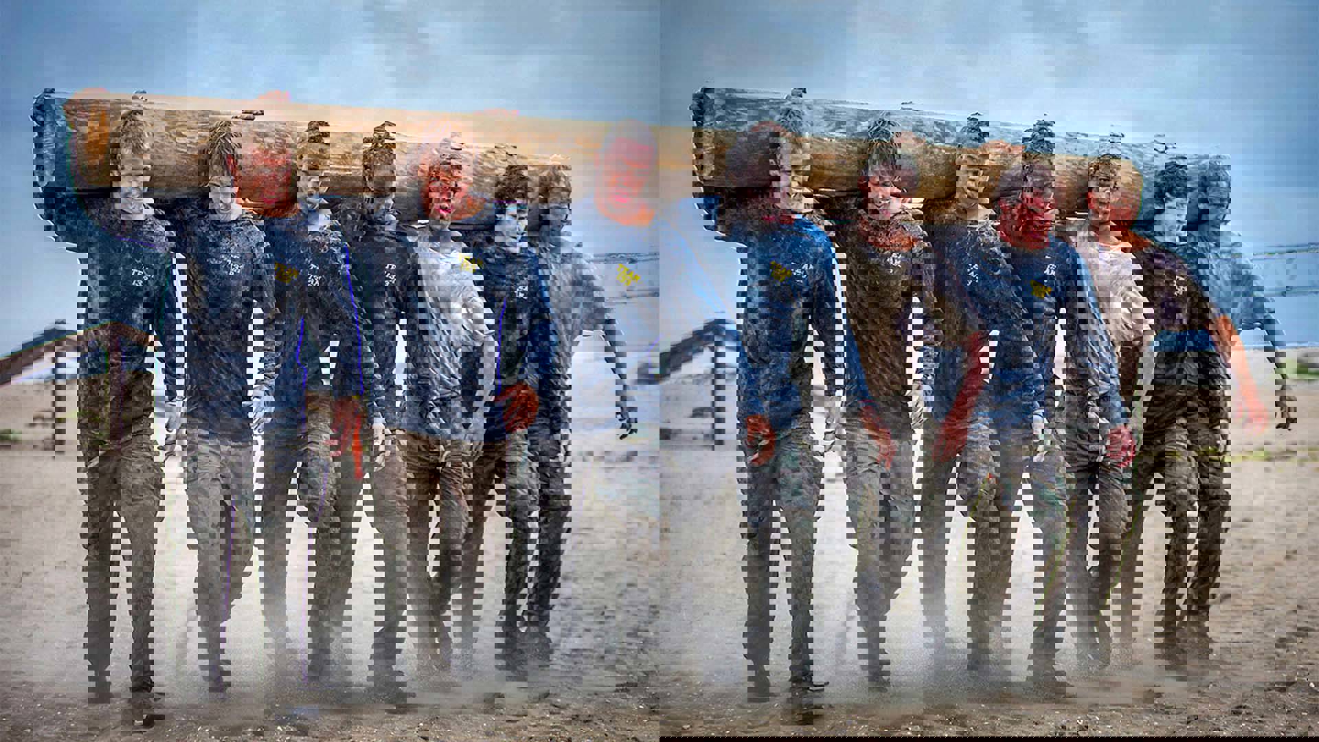 Seven men of various races holding a large wooden log in the sand.