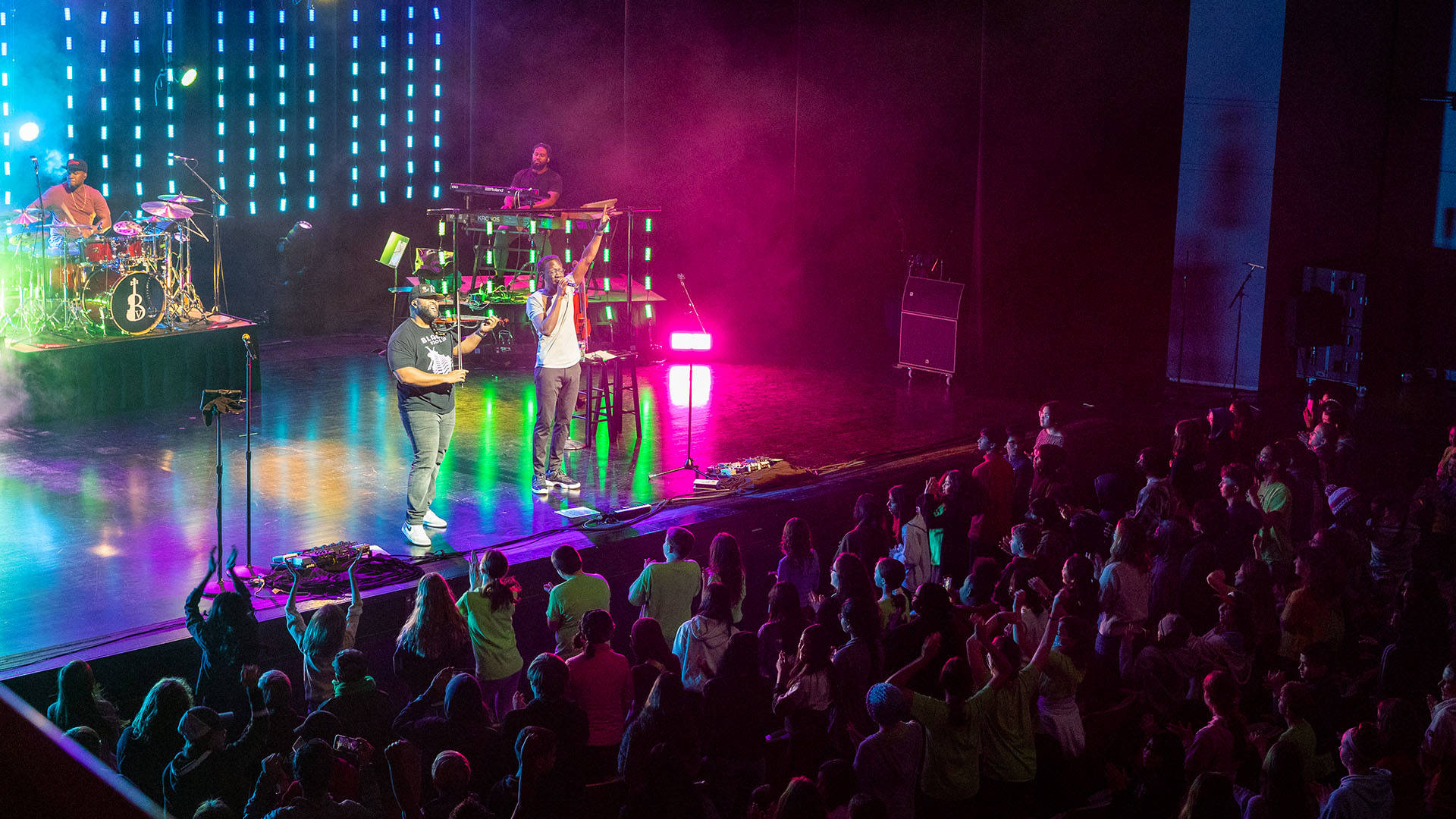 Black Violin performing on Overture Hall stage with a standing audience clapping. Four Black men playing drums, keys, violin and singing into a mic with multicolored stage lighting surrounding them.