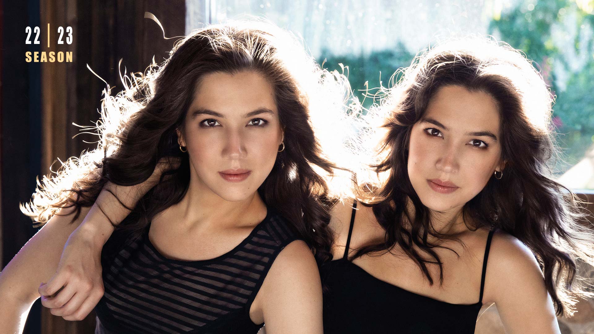 Banner text reads: 22 | 23 season. Two sisters hold each other and star intently into the camera. They both have brown eyes and long brown hair. They are wearing black tops.