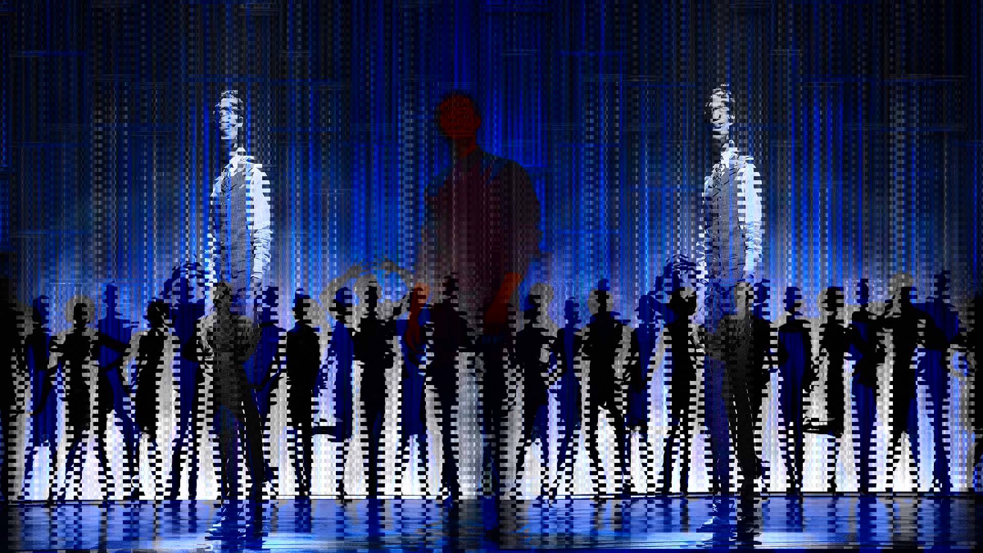 Noah Ricketts, a man with medium complexion wearing a vest and tie, performing on stage in "The Great Gatsby." The stage is light dark blue with silhouettes of background dancers posing against the light behind Noah.