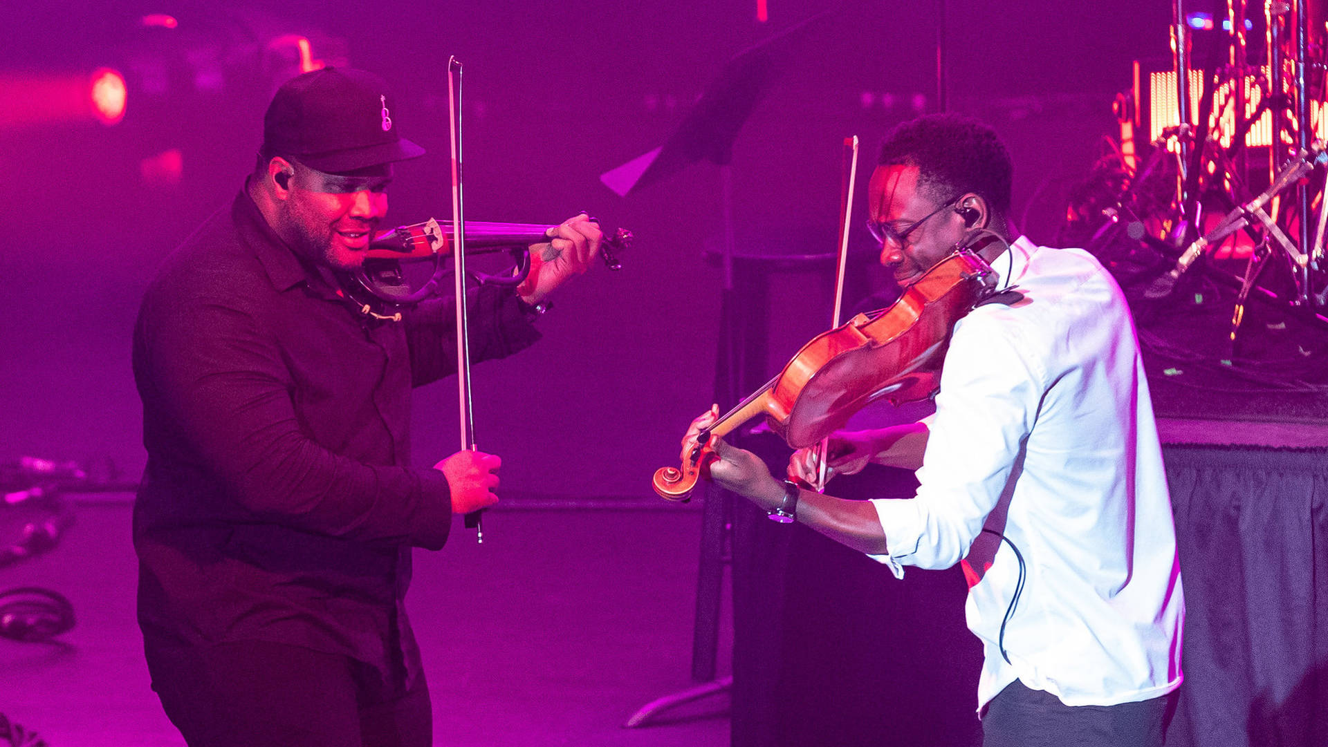 Two men in stylish clothing face eachother and play violins in reddish light. The one on the lef has a black baseball cap, dark complexion and dark clothing. The one on the right has short hair, dark complexion and a white shirt.
