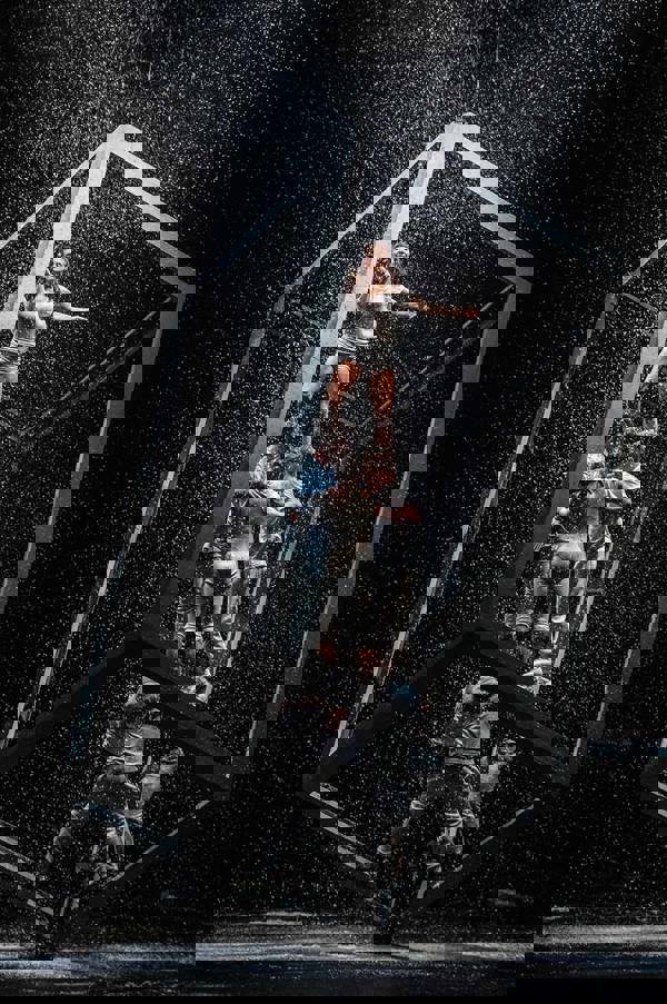 Seven dancers in light colored clothing. They are in a large framed box that is leaning on one corner. The dancers are standing on each other with one dancer at the top.