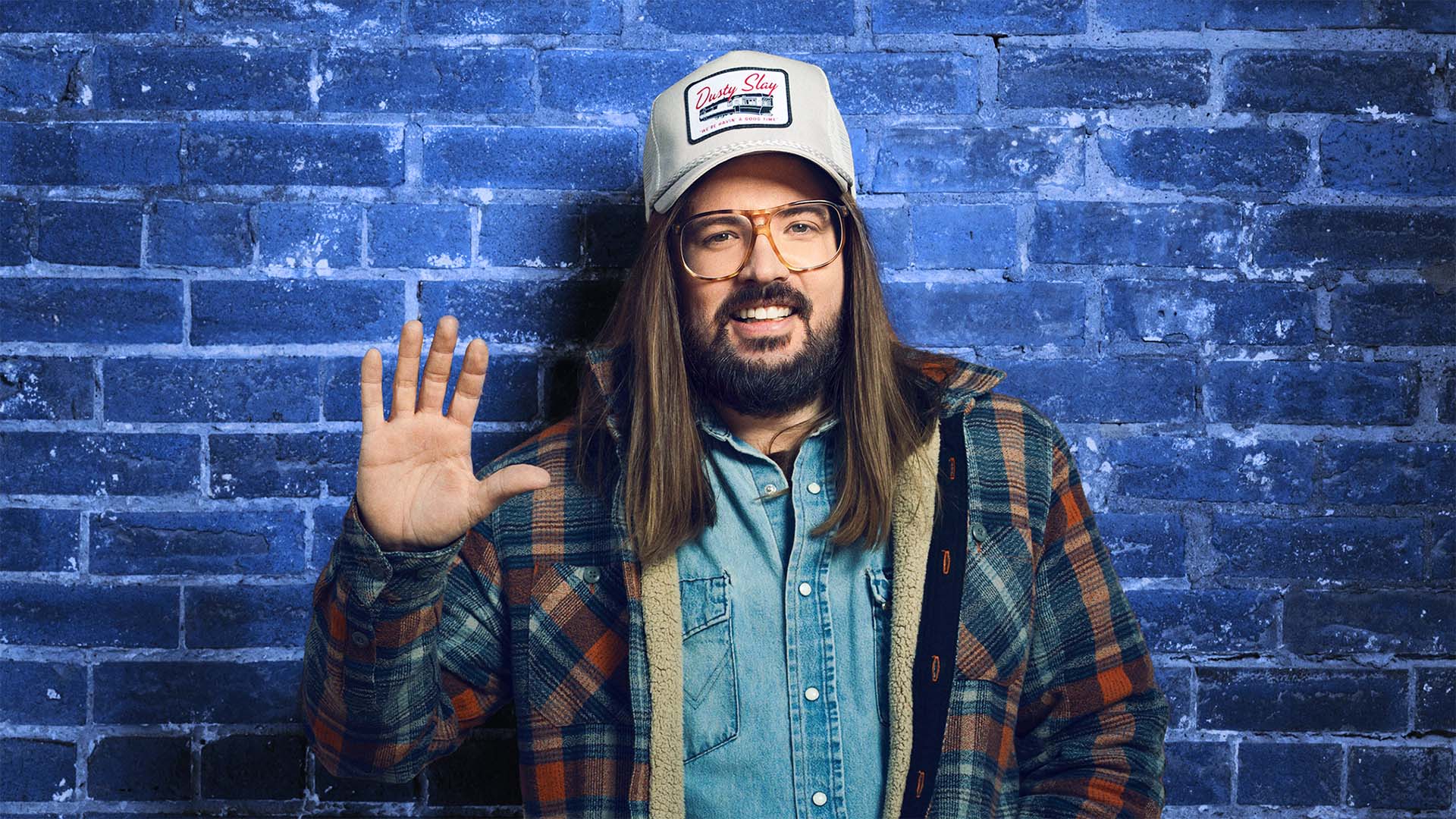 A white man with long brown hair, a trucker hat, glasses, and a flannel coat over jean shirt smiles and waves to the viewer. The background is a blue brick wall.