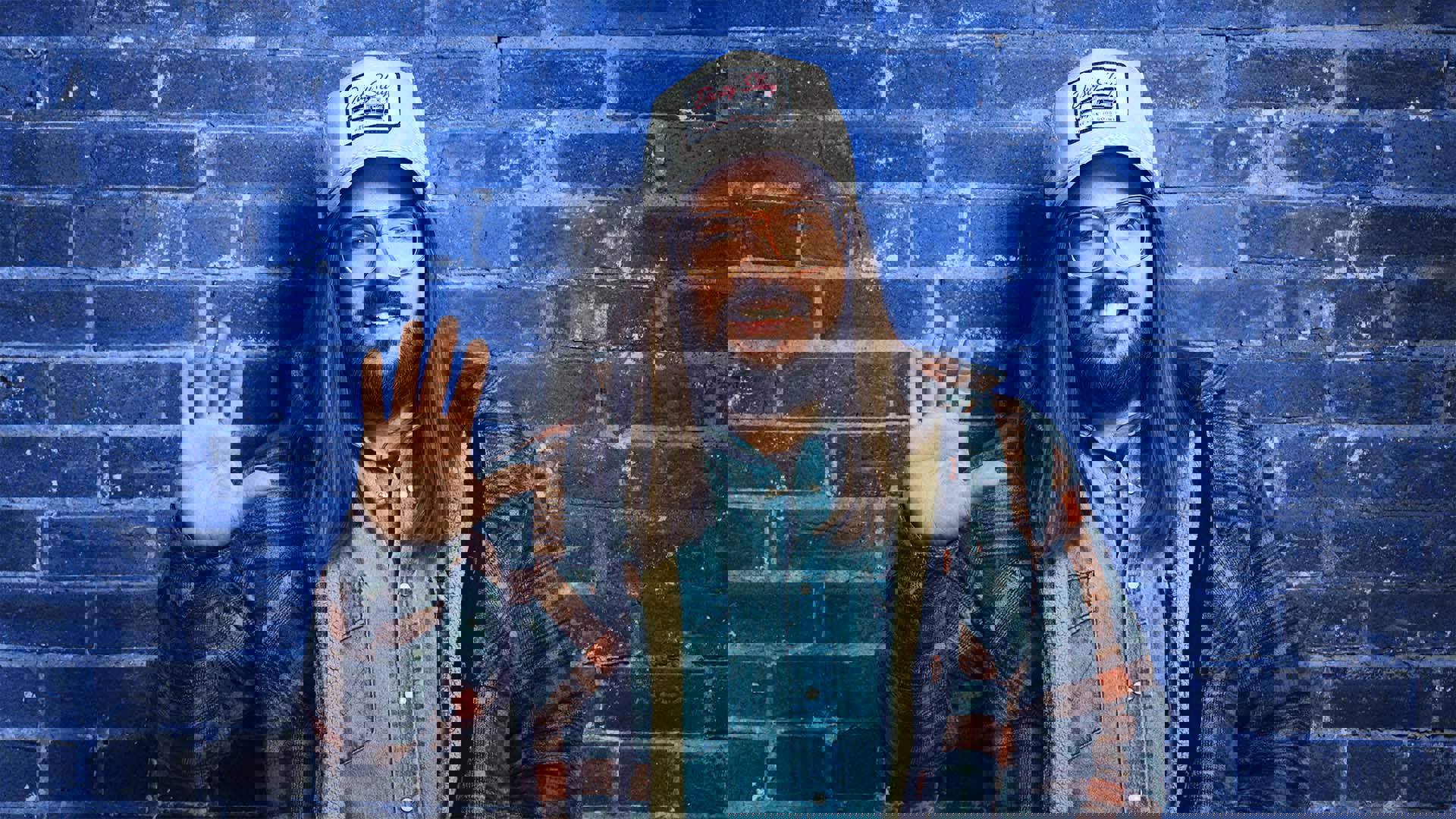 A white man with long brown hair, a trucker hat, glasses, and a flannel coat over jean shirt smiles and waves to the viewer. The background is a blue brick wall.