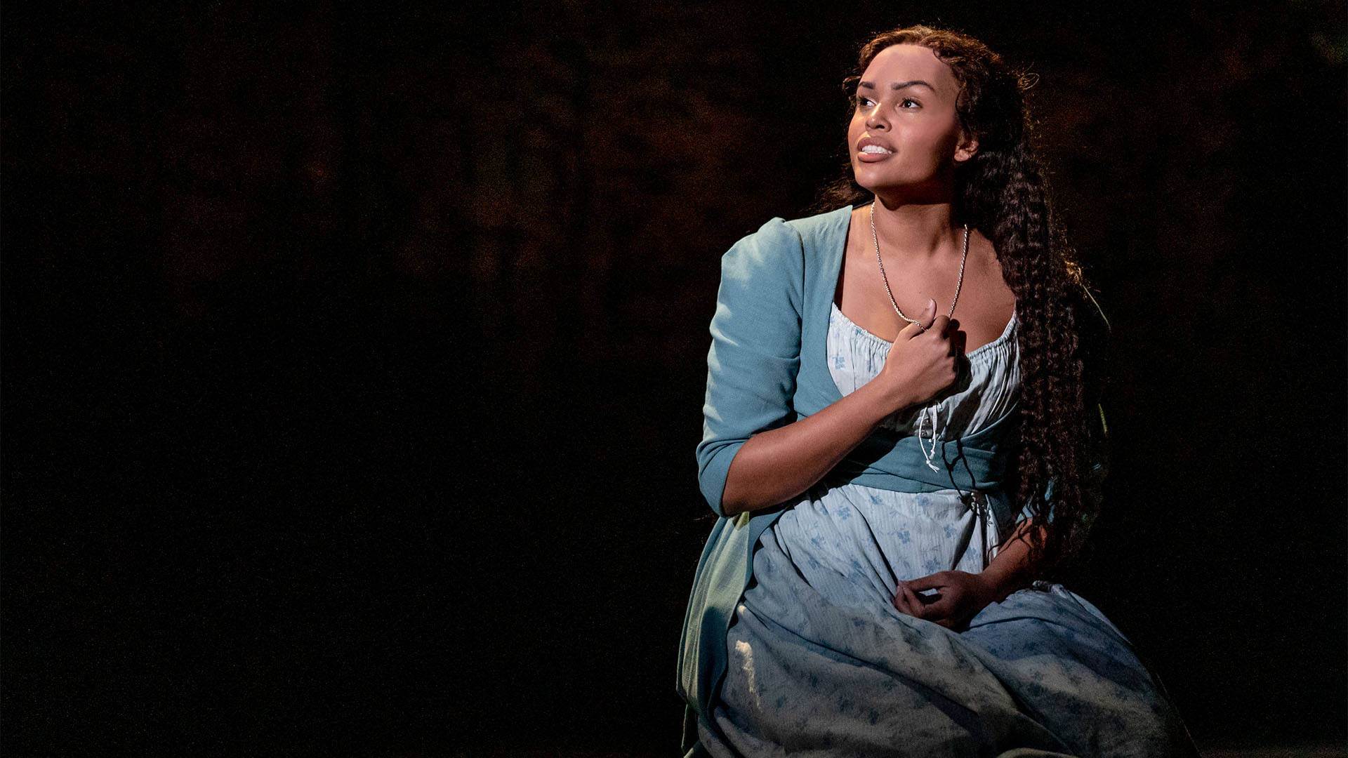 A woman in a blue dress with long dark hair singing and holding her fist against her chest.