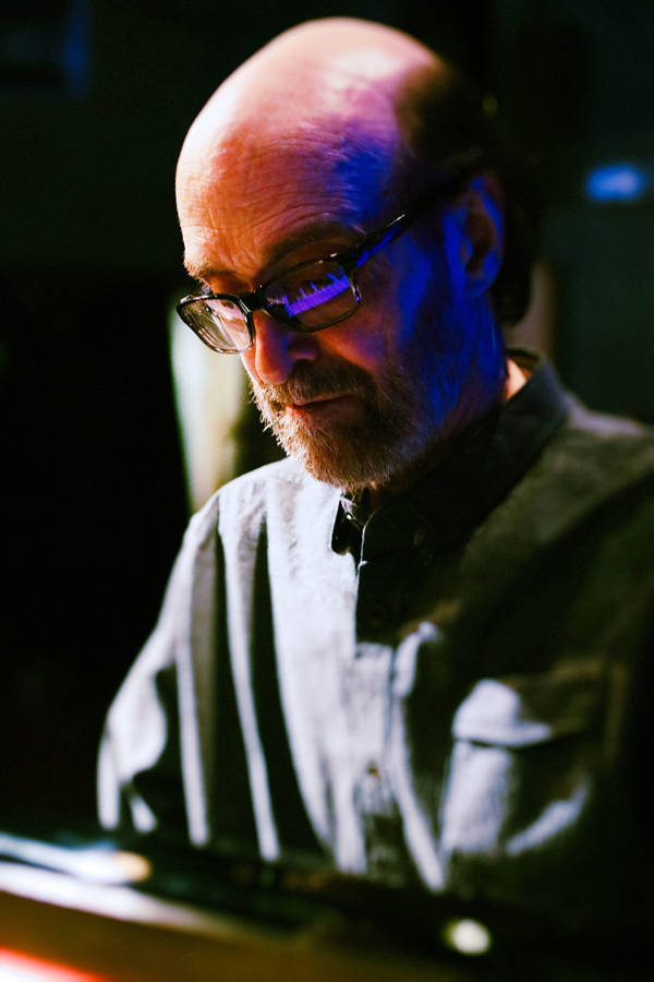 George Winston playing a piano. He is a caucasian man with a bald head, grey beard, and glasses