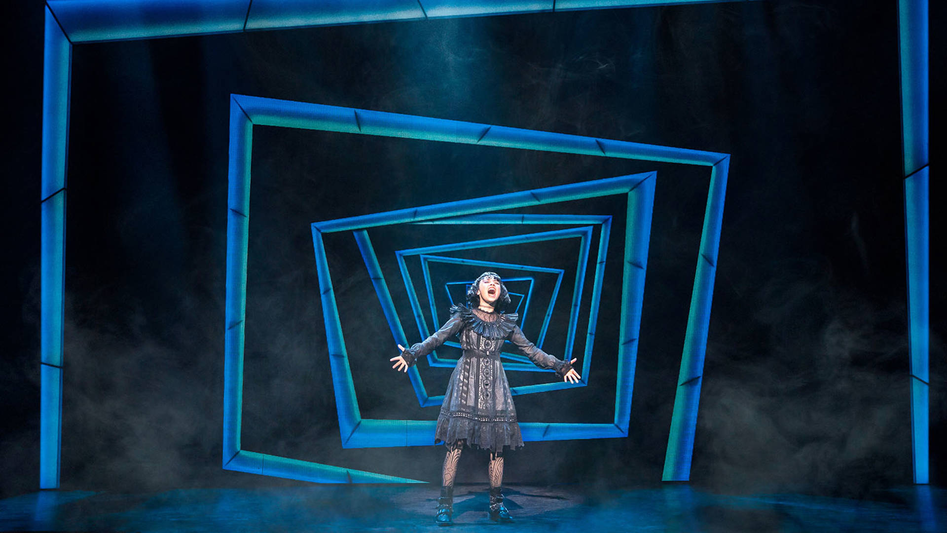 Lydia, a young woman in a black dress with black hair, performing on stage. Her arms are out wide.
