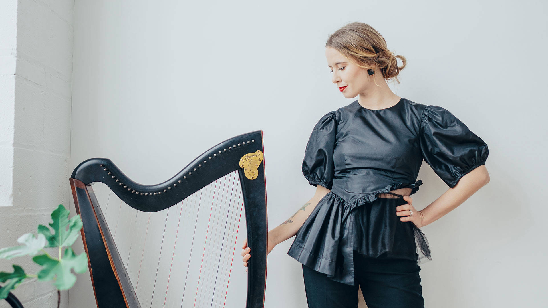 A caucasian woman with dark hair with blonde highlights pulled back and a dark outfit. She is standing next to a harp.