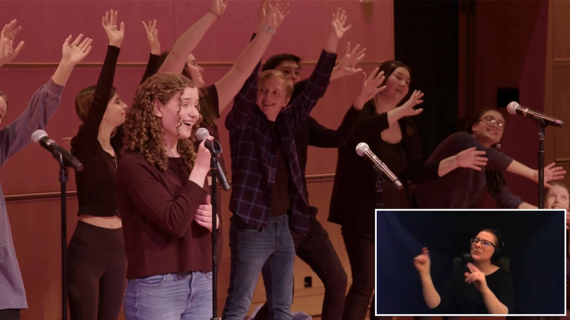 A screenshot of high school students singing and dancing. In the lower right corner an ASL interpreter is signing.