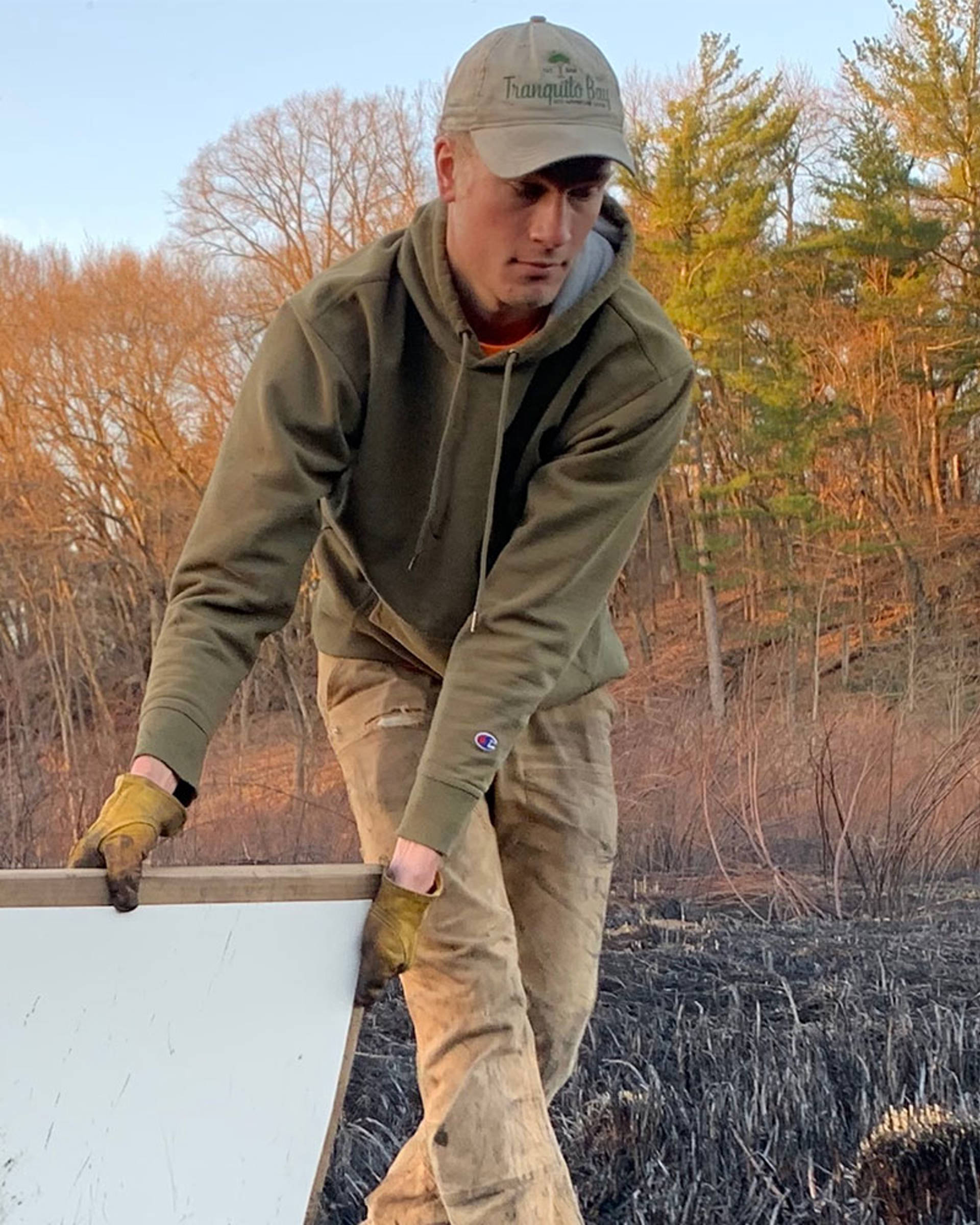 A young man with light complexion, gloves, a hat, and a hoodie, in a controlled burn field bracing a piece of wood.