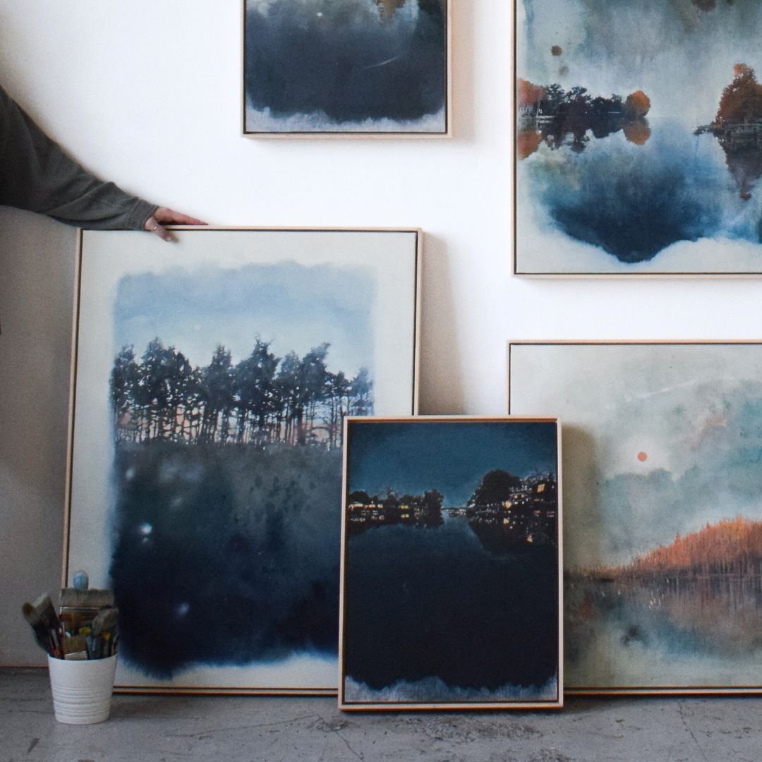 Multiple pieces of framed artwork leaning against a wall with blue, copper, and white tones. A hand is coming from the left and resting on one of the pieces.