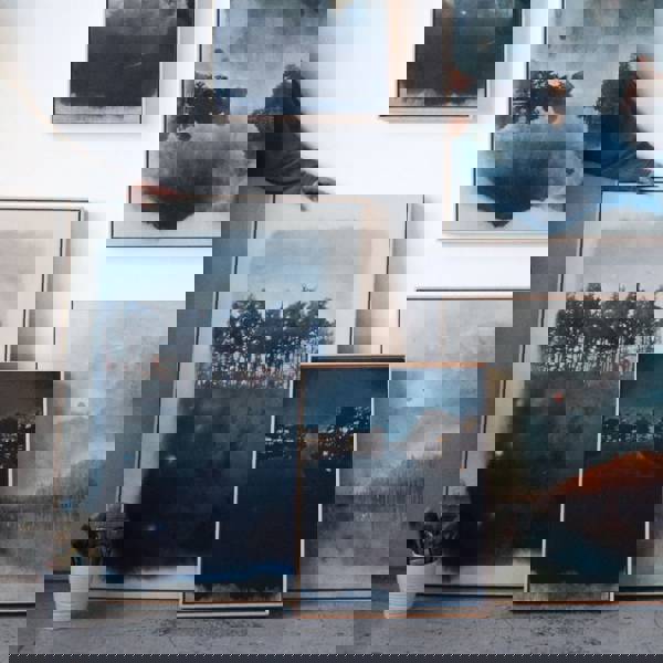 Multiple pieces of framed artwork leaning against a wall with blue, copper, and white tones. A hand is coming from the left and resting on one of the pieces.