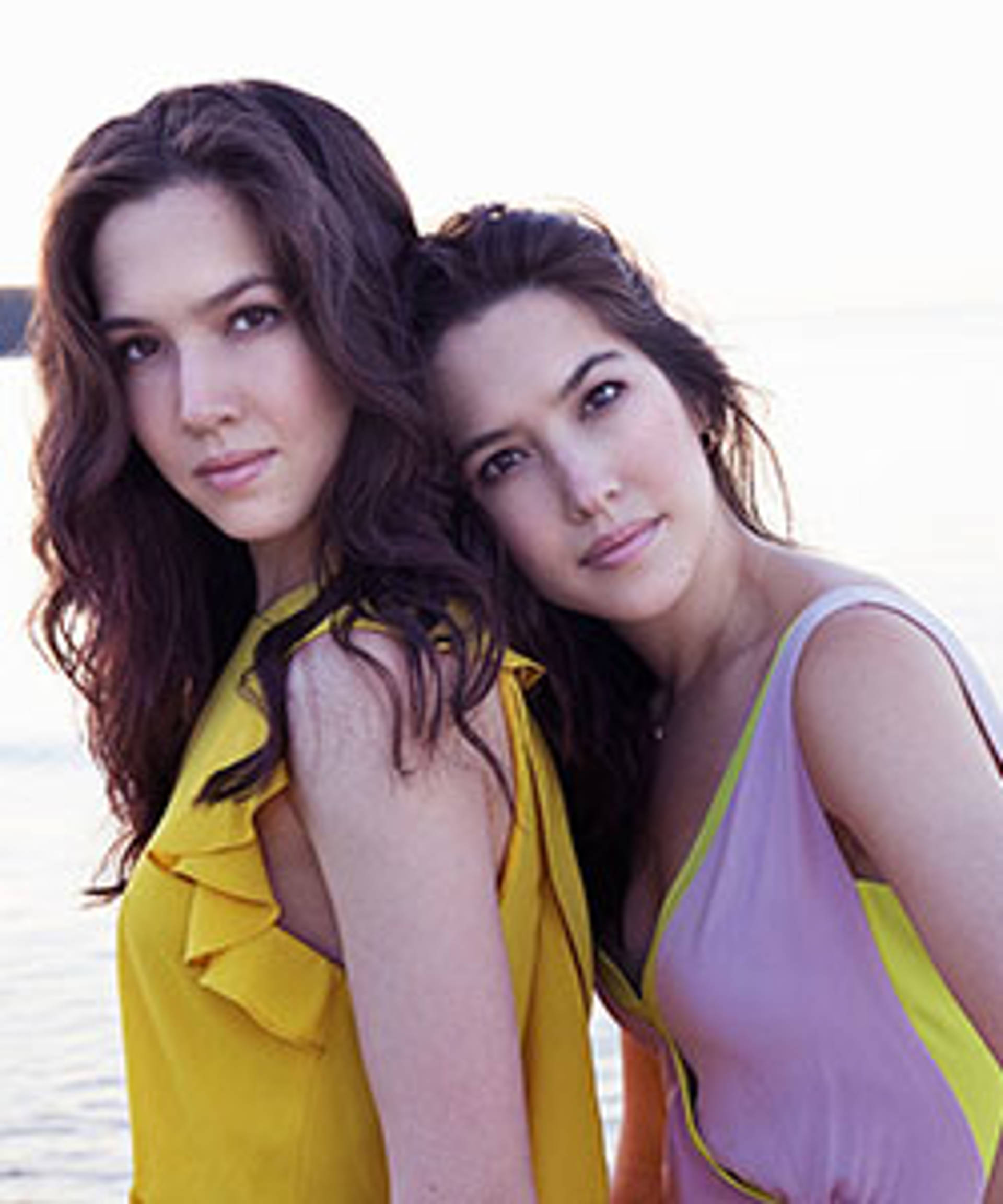 Image of two sisters with brown hair and dark eyes. one is wearing a yellow top and the other a pink top. One is leaning against the other.