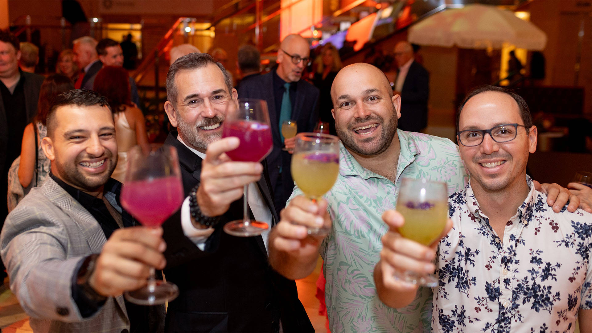 Four men with medium and light complexions smiling and raising cocktail glasses to the camera. They are wearing festive formal and casual shirts, two have suit jackets on.