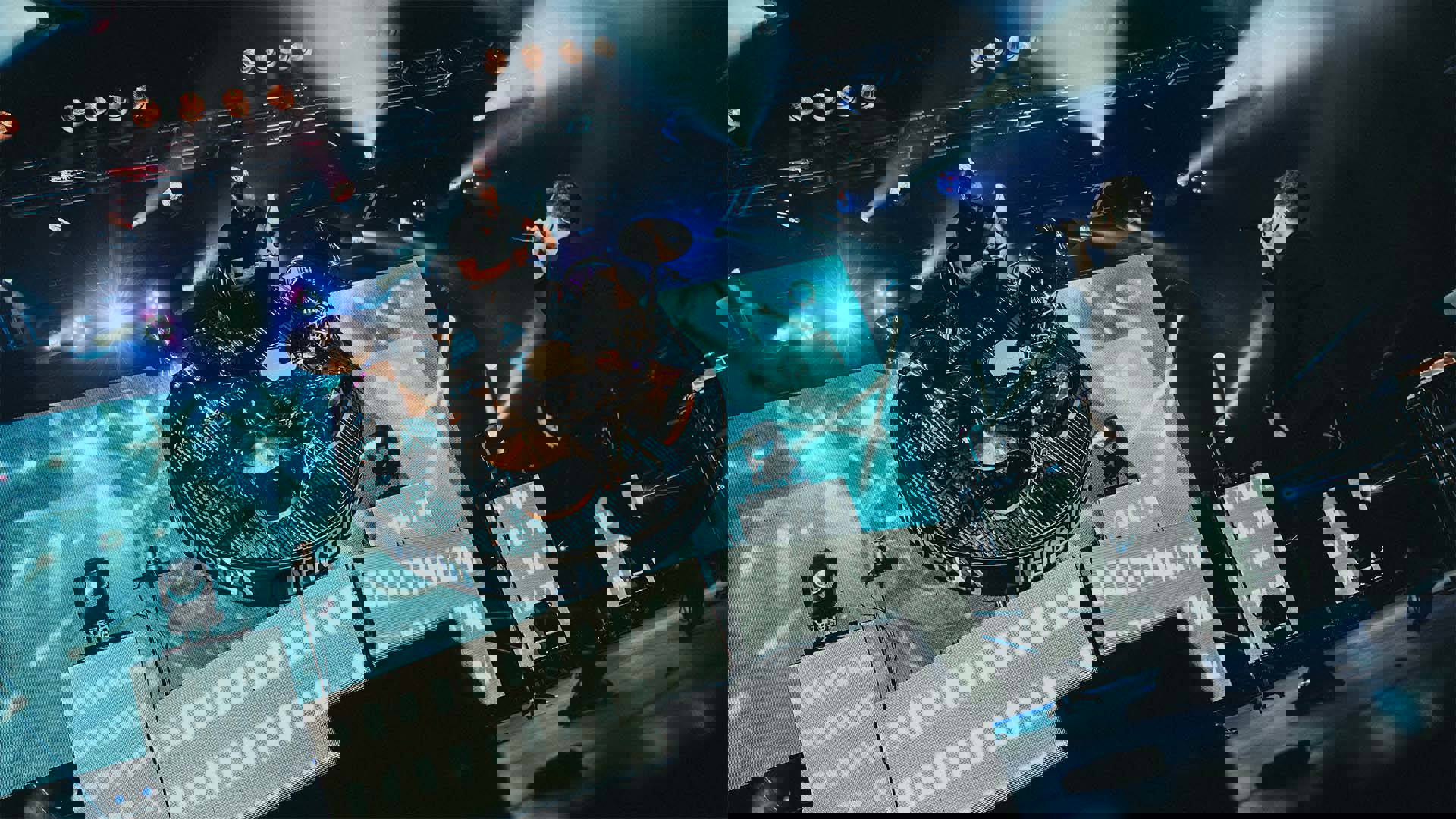 A man on a dramatically tilted rotating platform with drums playing a solo while another man looks on and sings into a handheld microphone.