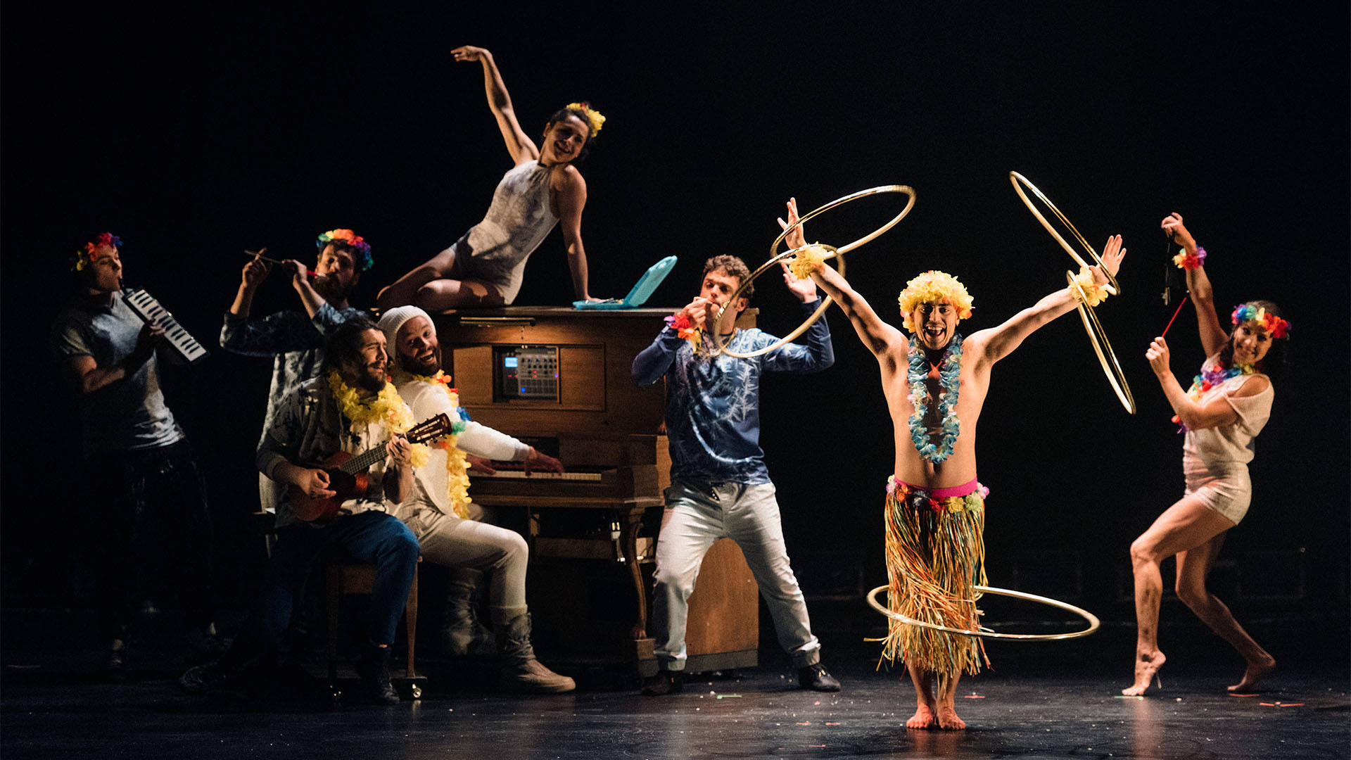 FLIP Fabrique performing on stage with a wooden piano. They are wearing flower boas and one is hula hooping in a straw skirt.