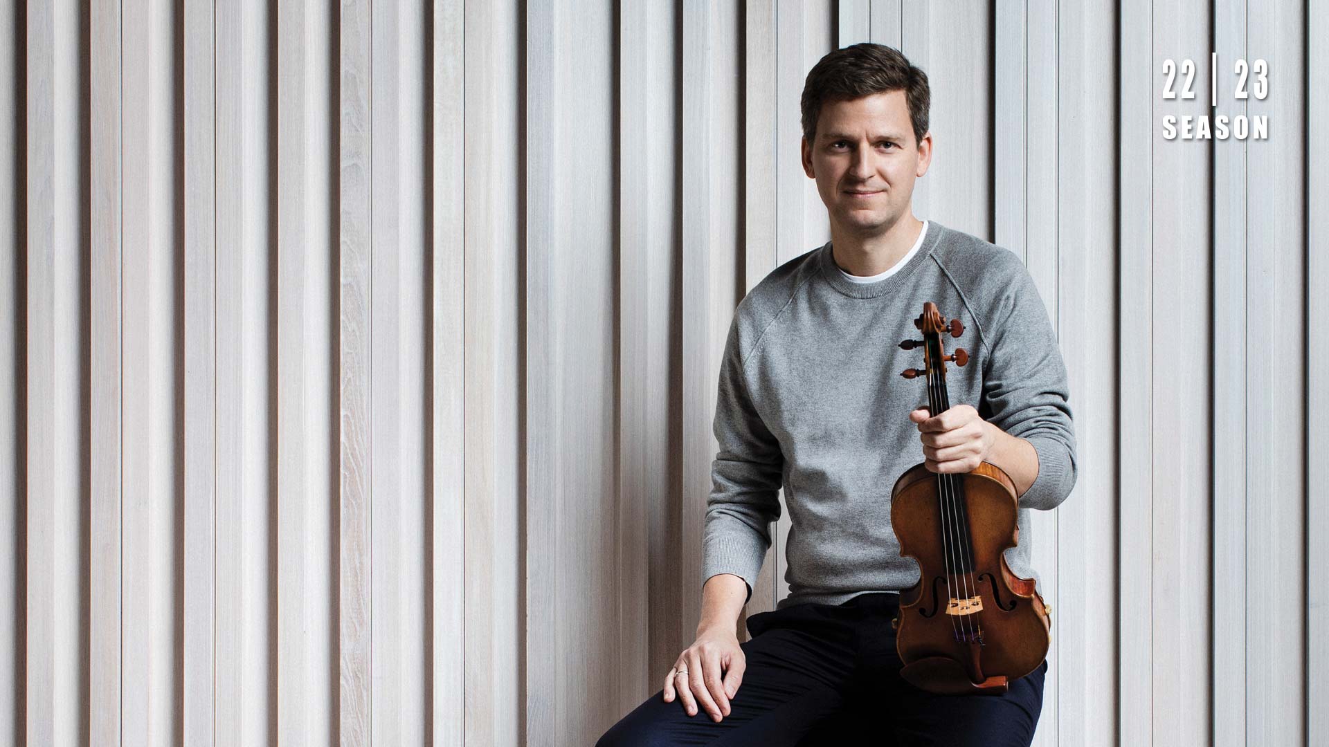 Banner text reads: 22 | 23 season. A caucasian man with short brown hair and a grey sweater sits holding a violin on his thigh.