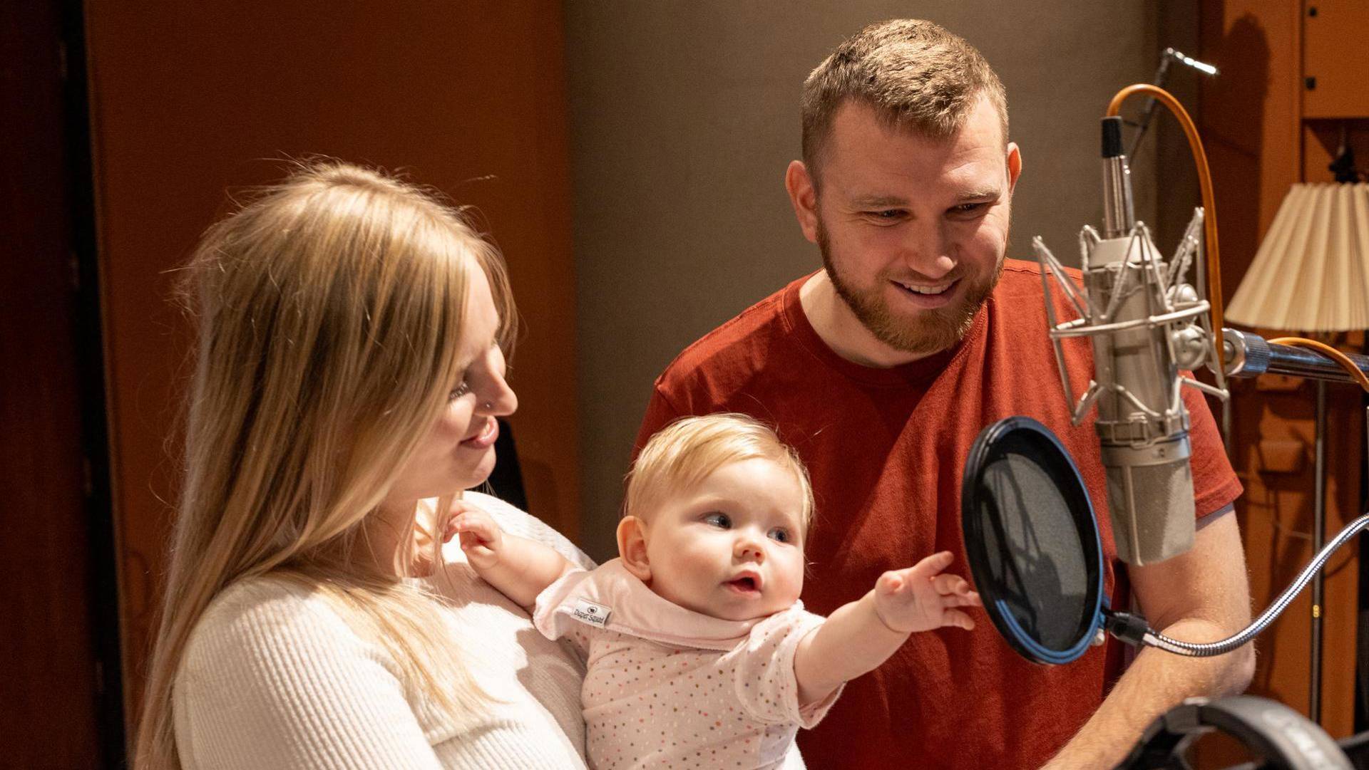 A mother and father with their baby looking at a high quality microphone. The baby girl is reaching for the microphone. They all have light complexion and lighter hair.