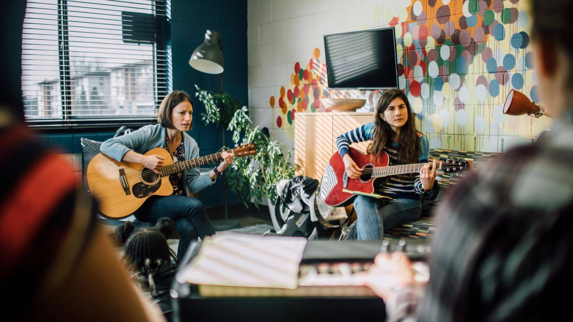 Two woman sitting playing acoustic guitar together. A colorful brick wall and window are behind them. In a foreground another musicians blurry hand is playing a keyboard.