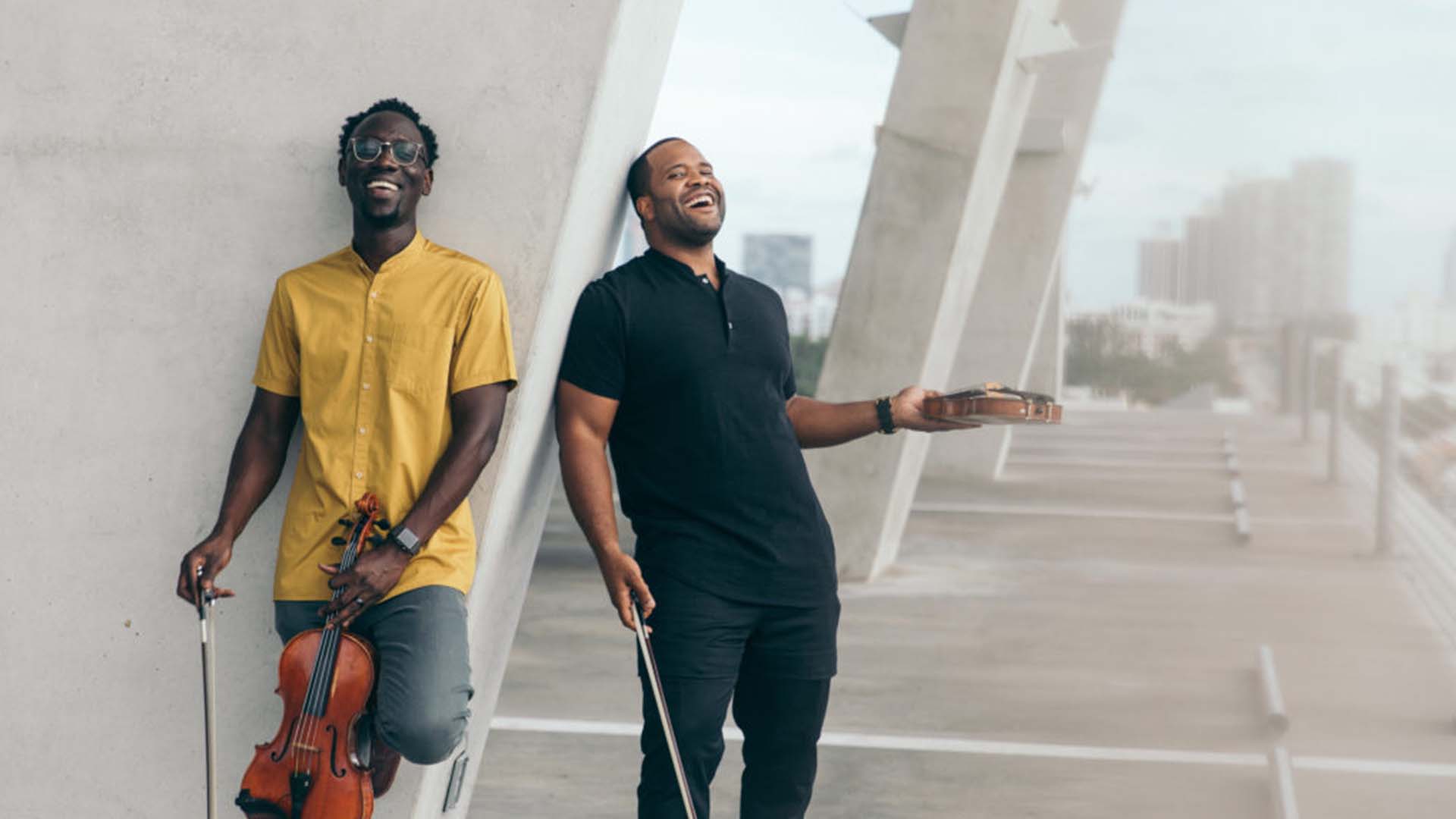 The two members of Black Violin lean against a concrete slab and laugh. They are both holding a violin and a bow. One has a gold button up shirt and the other has a black polo. They both have short hair