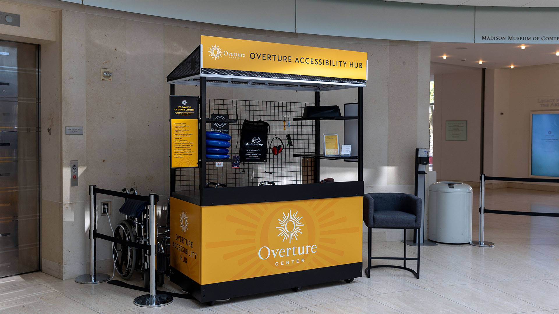 A black metal kiosk with yellow Overture branding in Rotunda lobby. The top banner reads: "Overture Accessibility Hub." There are various items on display, including sensory-friendly tools like wobble seats and headphones.