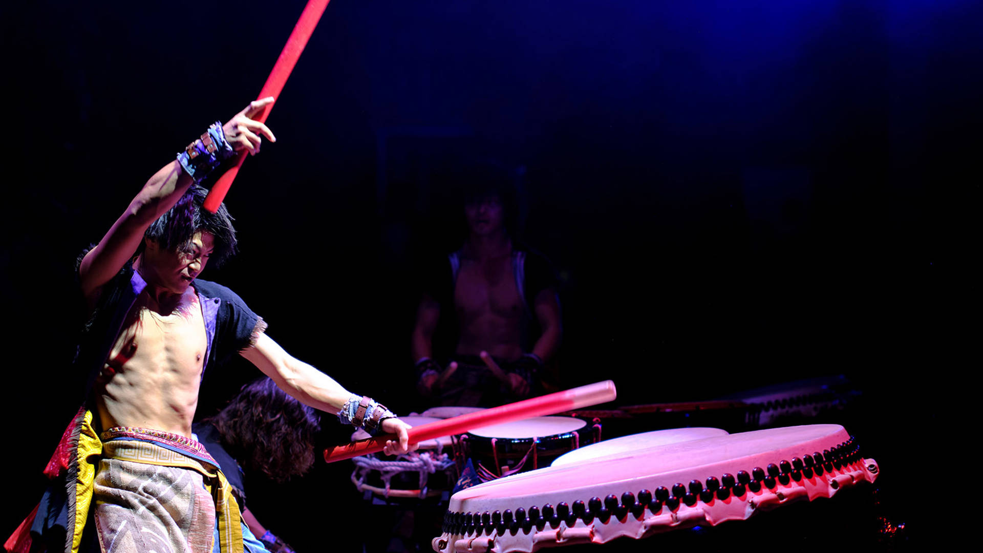 A YAMATO drummer holding two large red drumsticks in mid strike of a large drum.