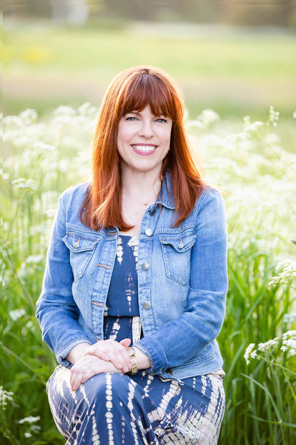 A white woman with reddish-brown, shoulder-length hair. She is sitting in a grassy field, wearing a jean jacket and a blue dress.