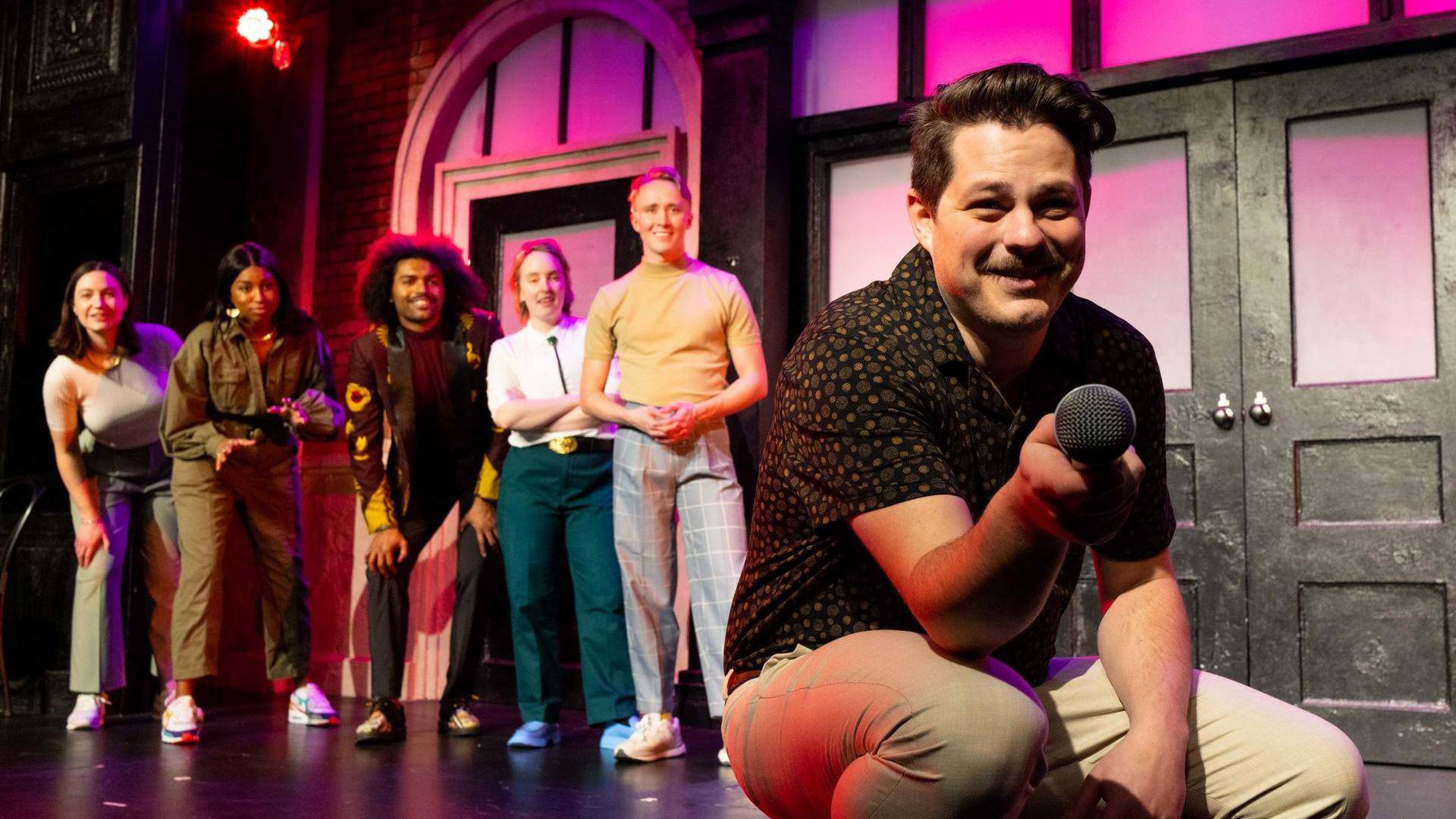 A caucasian person with short brown hair and a mustache holds a mic out to the audience/camera. Behind them are five people also on stage smiling.
