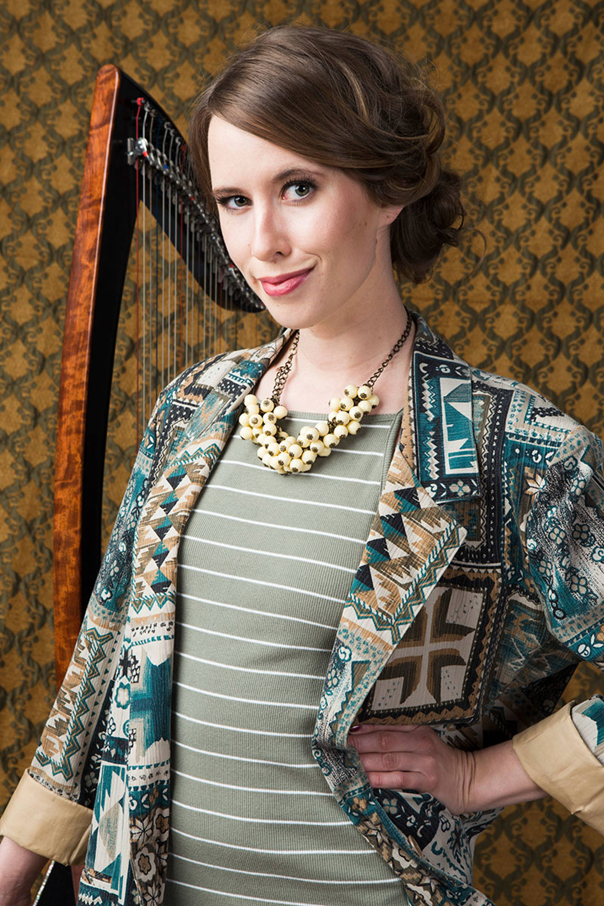 headshot of caucasian woman with dark medium length hair and brown eyes. A harp is standing behind her. She has a green top with white stripes and a colorful jacket.