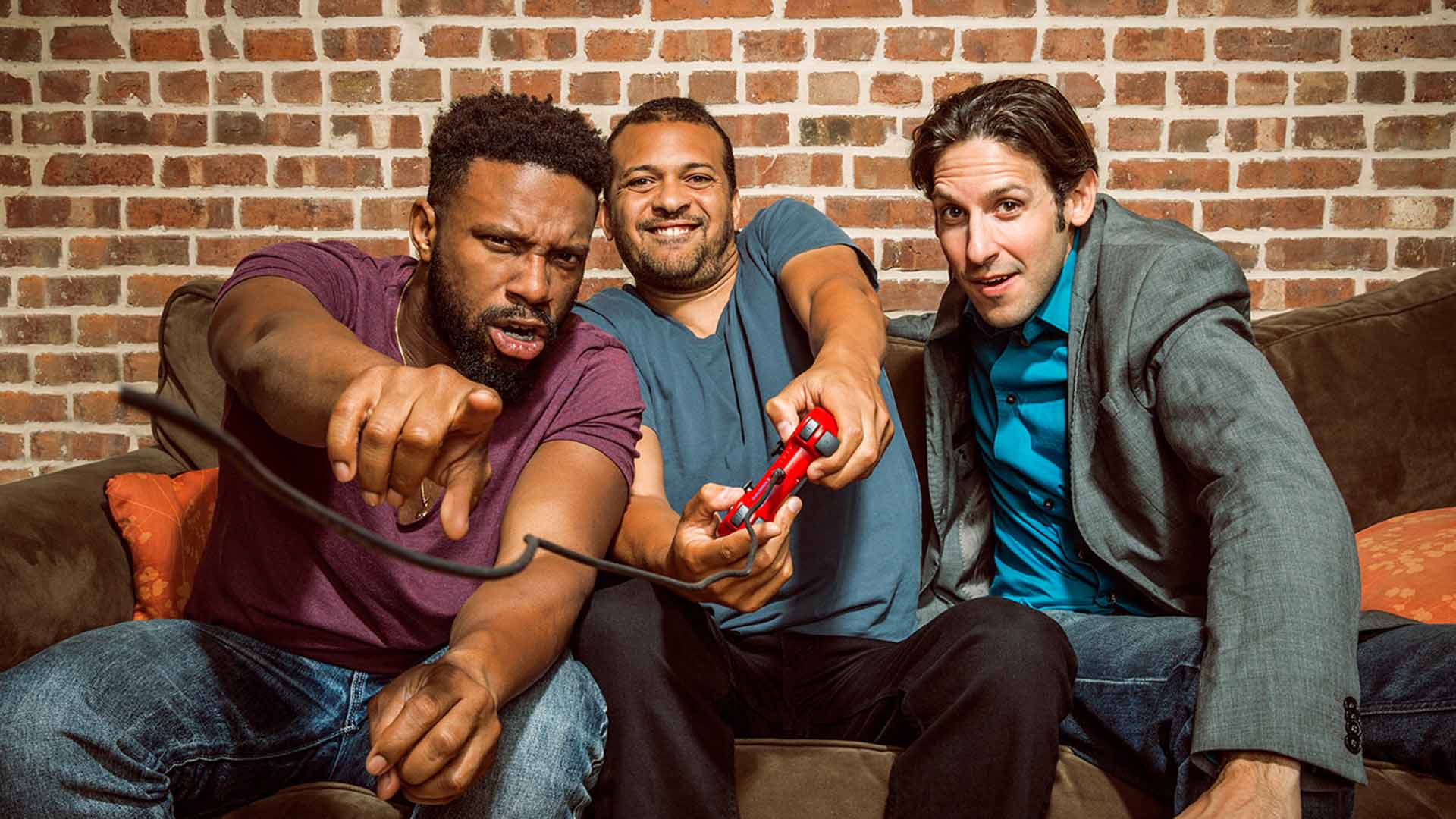 The Mayhem Poets are sitting on a couch actively playing video games. Two are African American men with short dark hair and purple and blue t-shirts. The third is a caucasian man with a suit jacket and medium brown hair. 