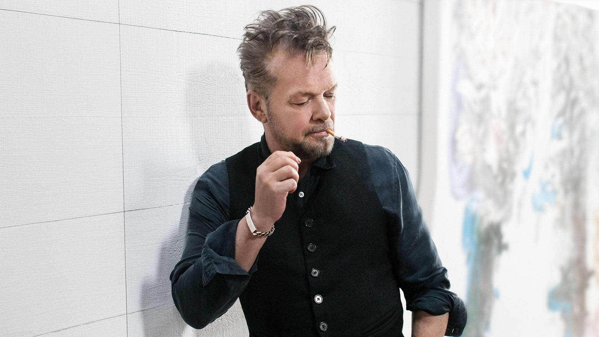 An image of John Mellencamp - a middleaged man with light complexion and short messy grey hair and wearing a black buttonup shirt.He is leaning against a beige wall and has a cigarette in this mouth.