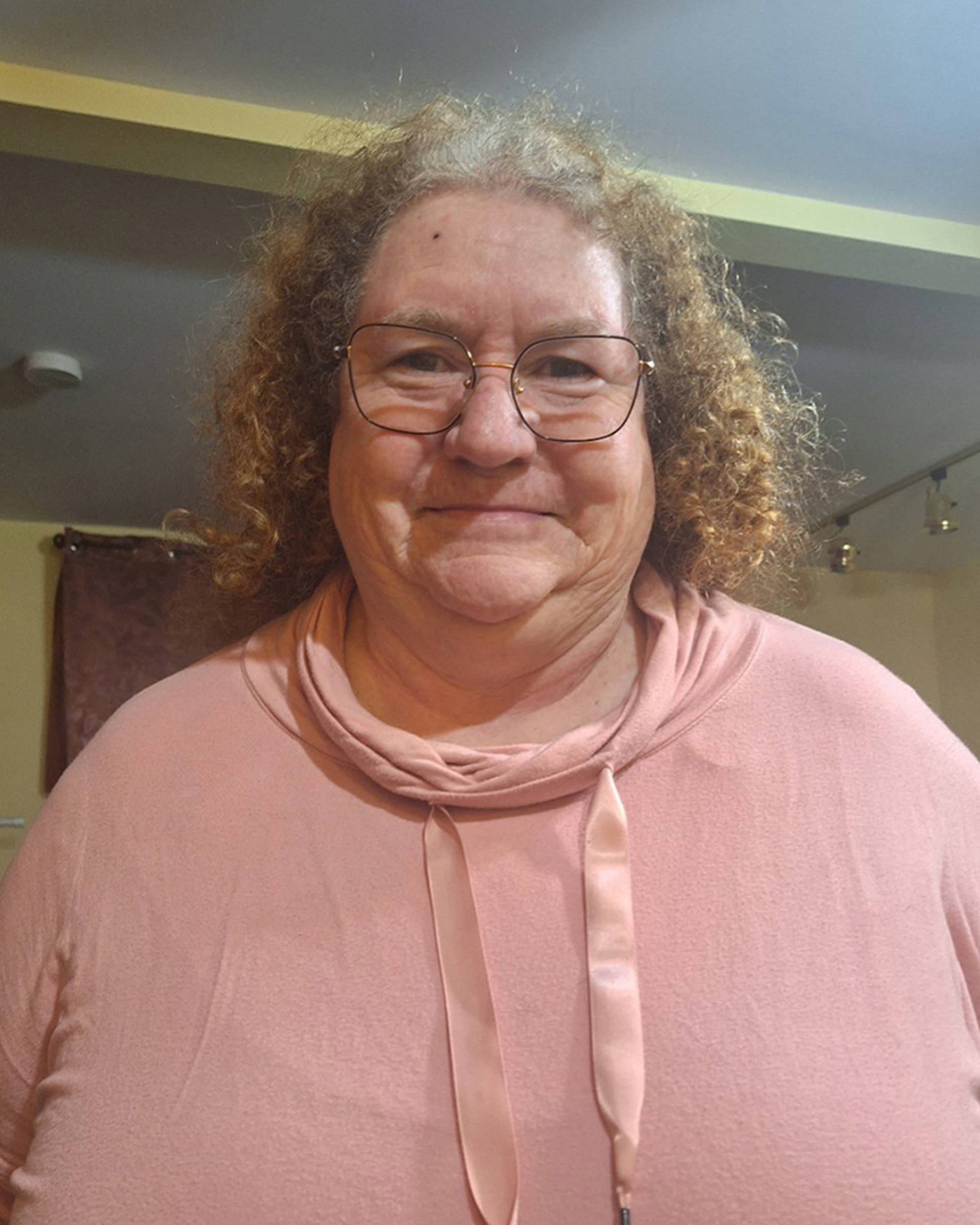 Headhot of a woman with light complexion, curly brown hair and glasses. She has a light pink top on.