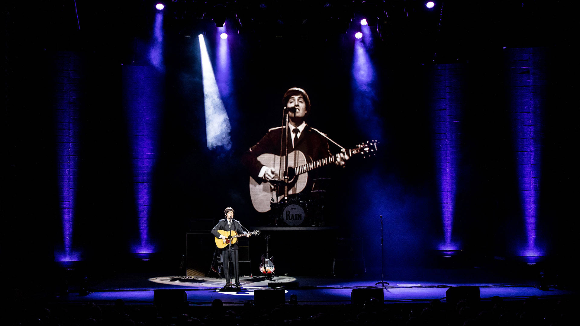 A Paul McCartney impersonator wearing a dark suit and holding an acoustic guitar. He has dark hair and is singing the song Yesterday on a dark stage.