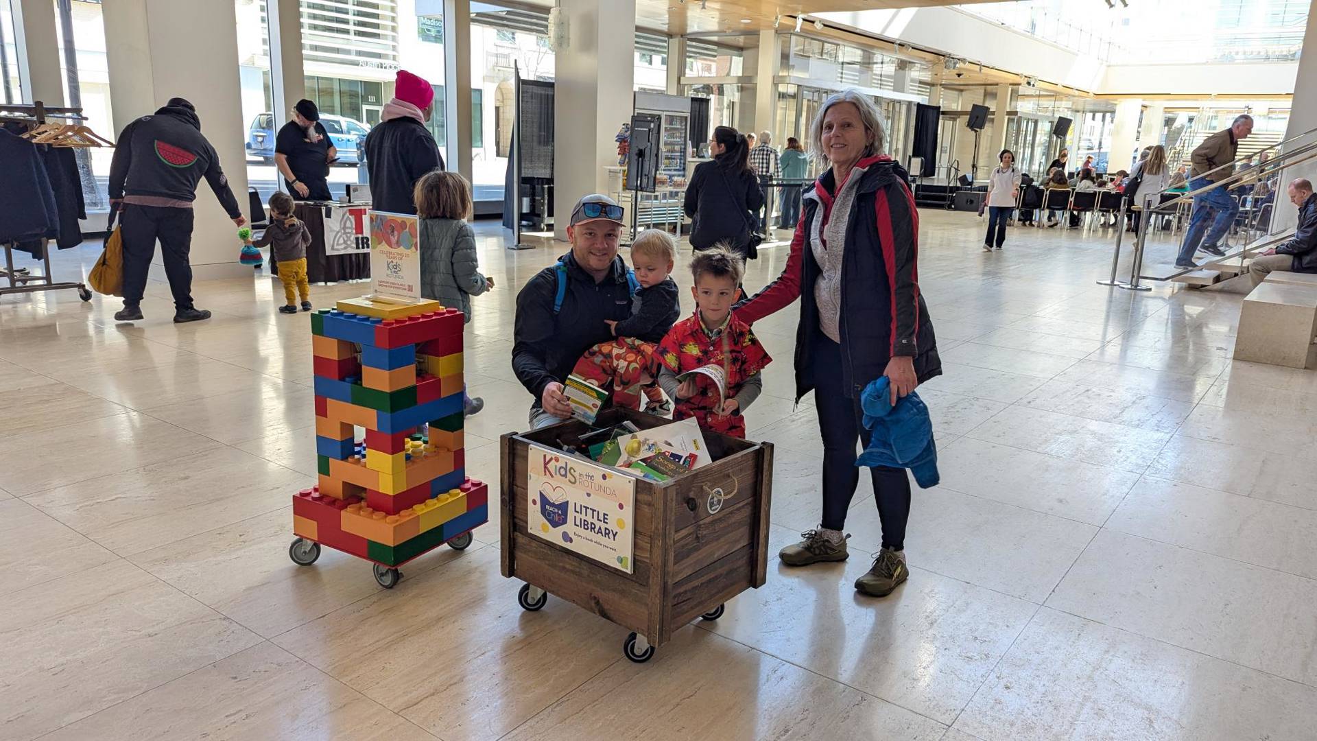 Two adults and two children smiling in Overture Hall Lobby  with a crate full of books.
