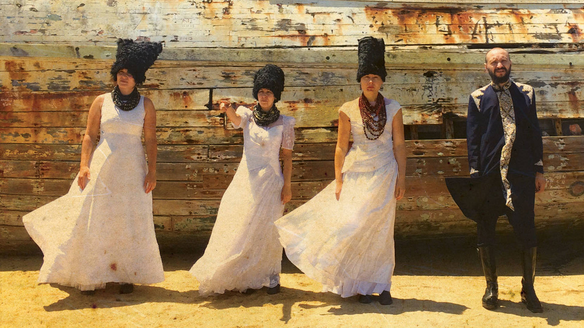 Four people with light complexion stand in a dusty beige outdoor scene in front of a wooden background. The three women are wearing white dresses with plush black headwear. The man is wearing a dark ukrainian top with ornamentation.