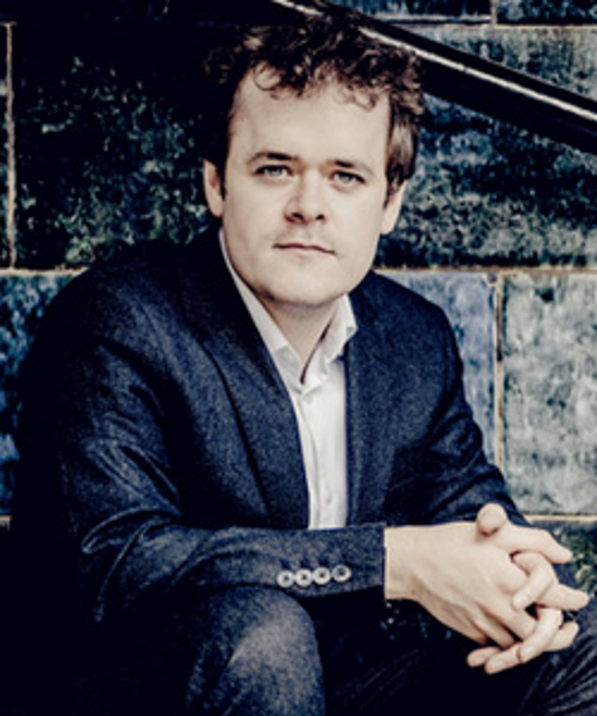 Headshot of a caucasian man with blue eyes messy brown hair. He is wearing a jacket and sitting with his hands clasped together