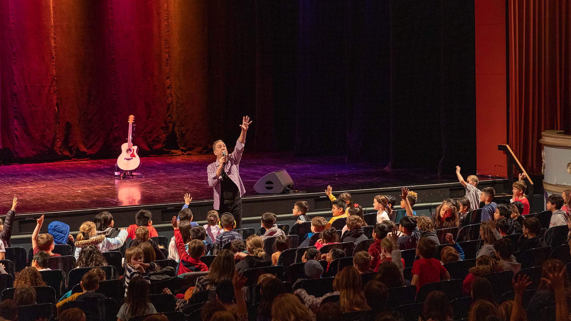 A man with medium complexion stands at the front of the Capitol Theater stage holding a mic and his other hand stretch up high. An audience of young students reach their hands up matching him in excitement.