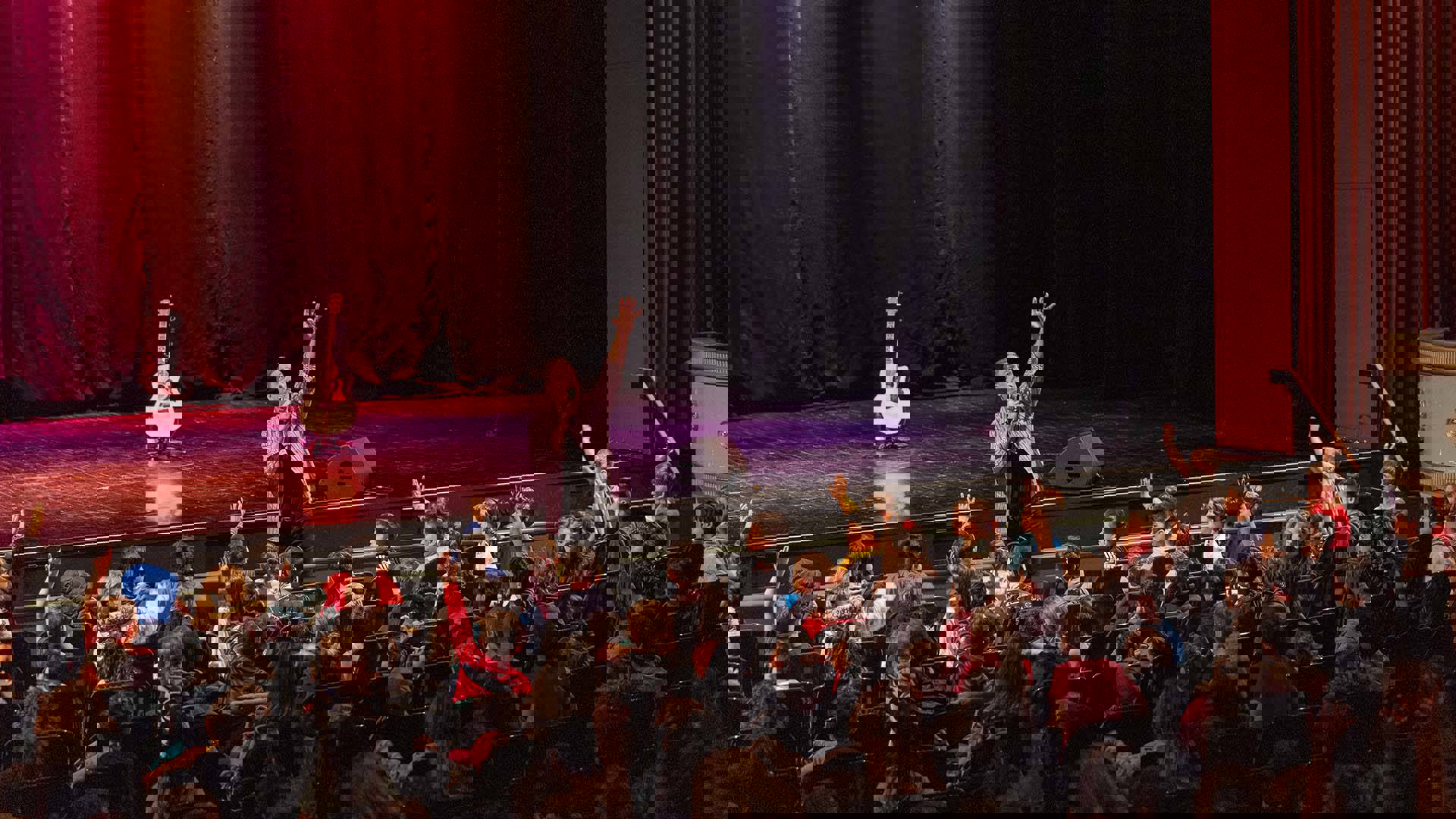 A man with medium complexion stands at the front of the Capitol Theater stage holding a mic and his other hand stretch up high. An audience of young students reach their hands up matching him in excitement.