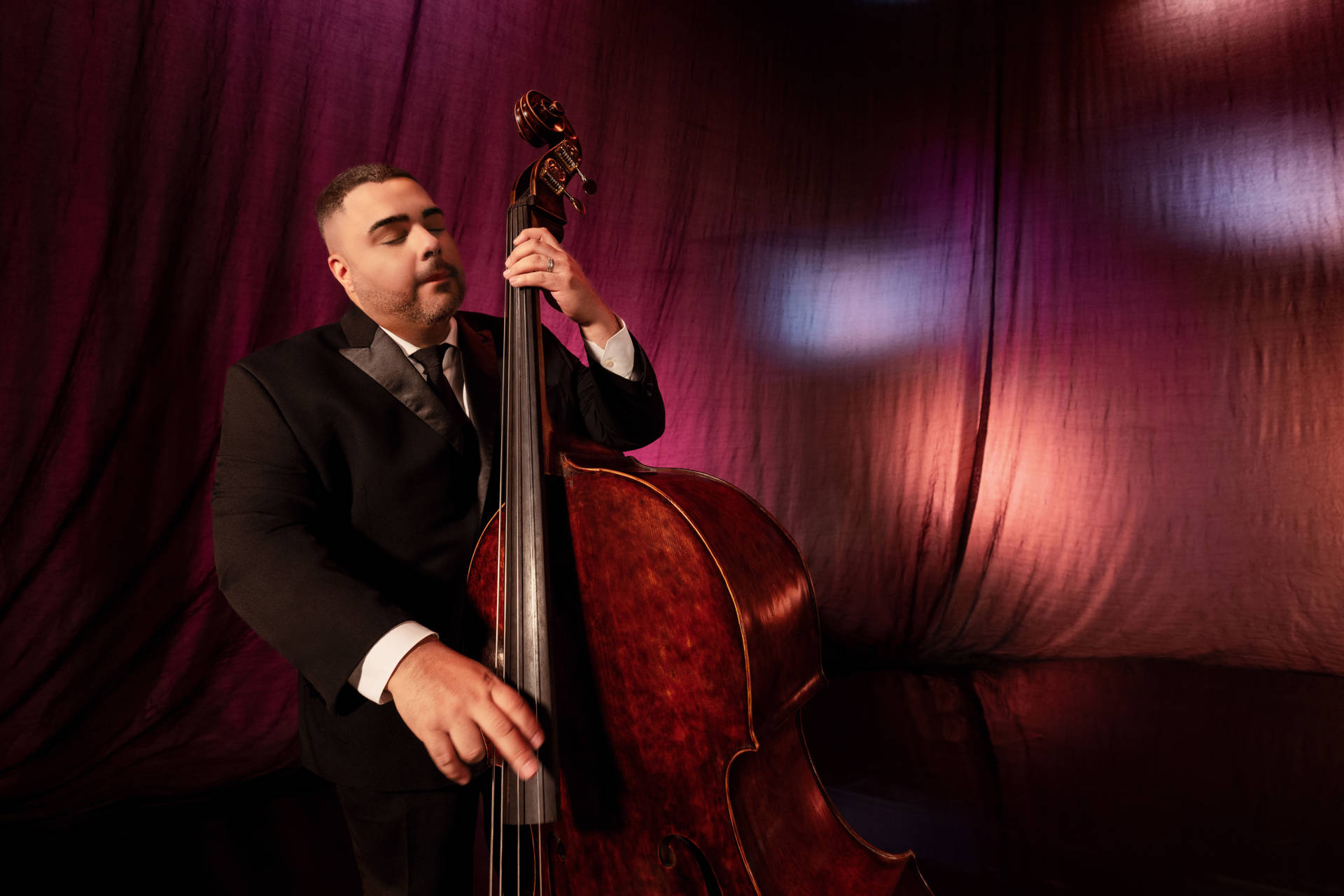 A man with medium complexion and dark hair plays a double bass in front of a pink stage curtain.
