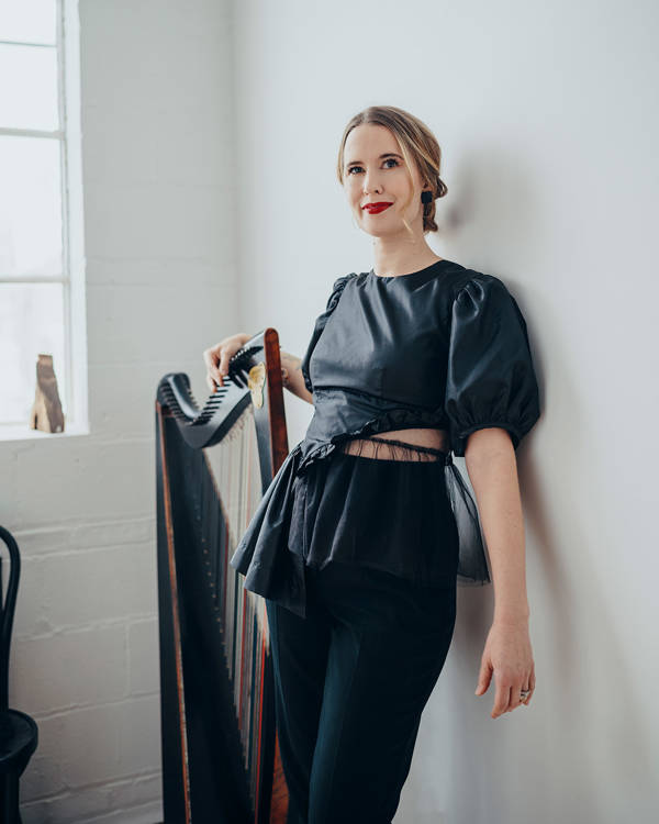 A caucasian woman with dark hair with blonde highlights pulled back and brown eyes standing against a white wall with a harp.