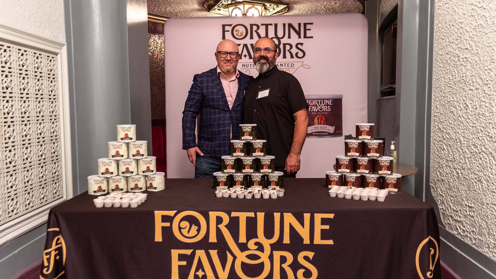 Two men stand together for a photo. they are both light complexion, wearing glasses, and bald. In front of them is a table with piled cans of food. The table and a banner behind them read "Fortune Favors" 